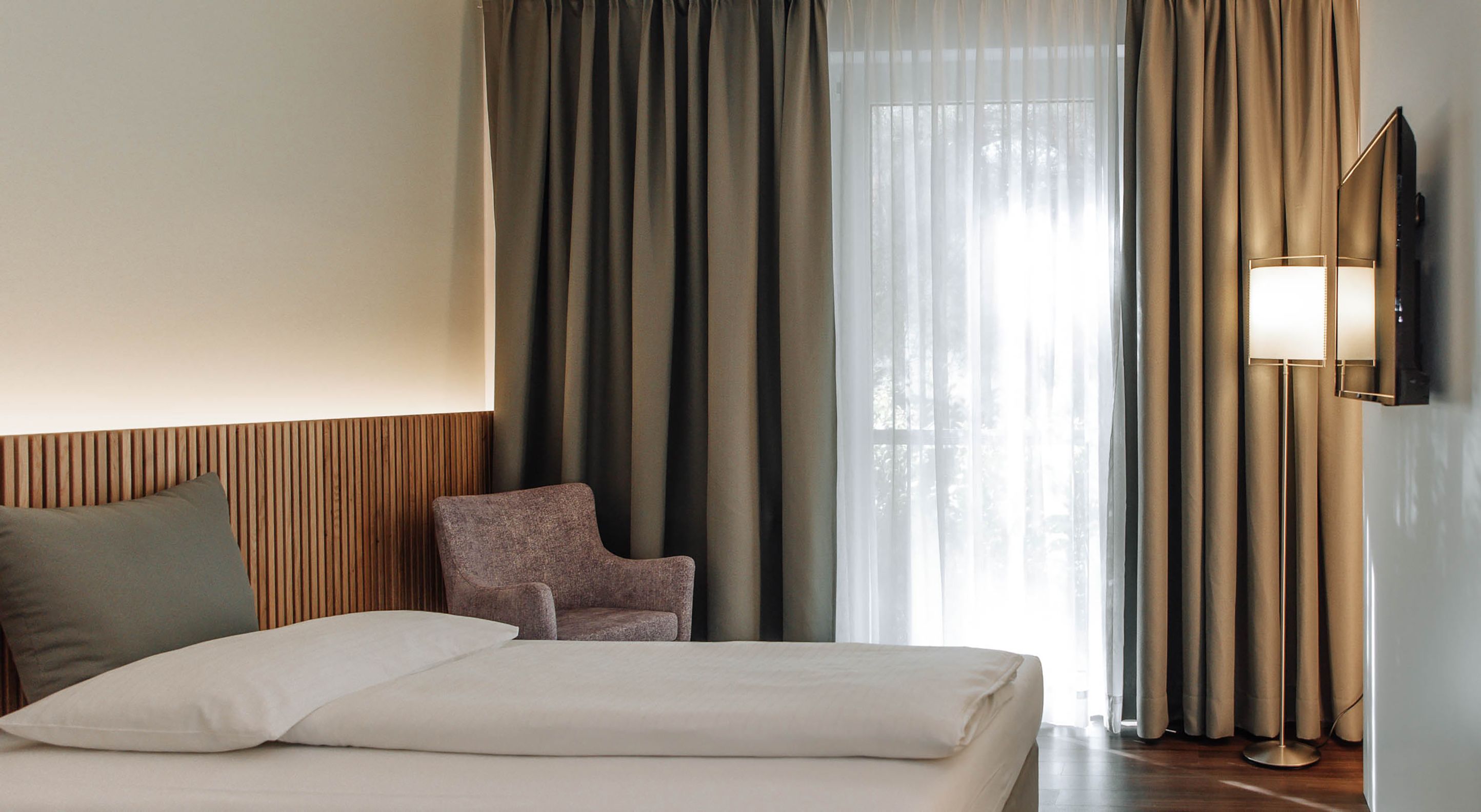 Modernes Hotelzimmer mit Bett, Sessel und Vorhängen.