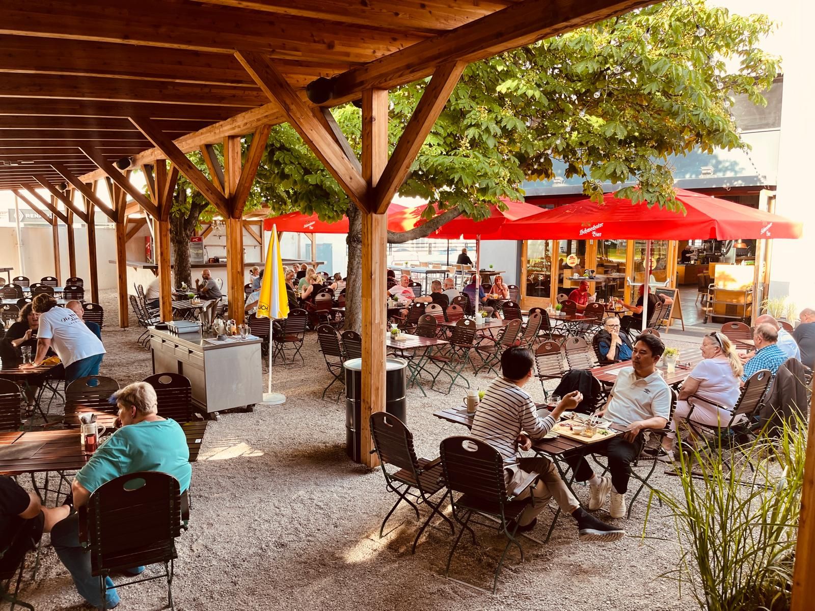 Ein Biergarten mit Holzdach, roten Sonnenschirmen und vielen Menschen an Tischen.