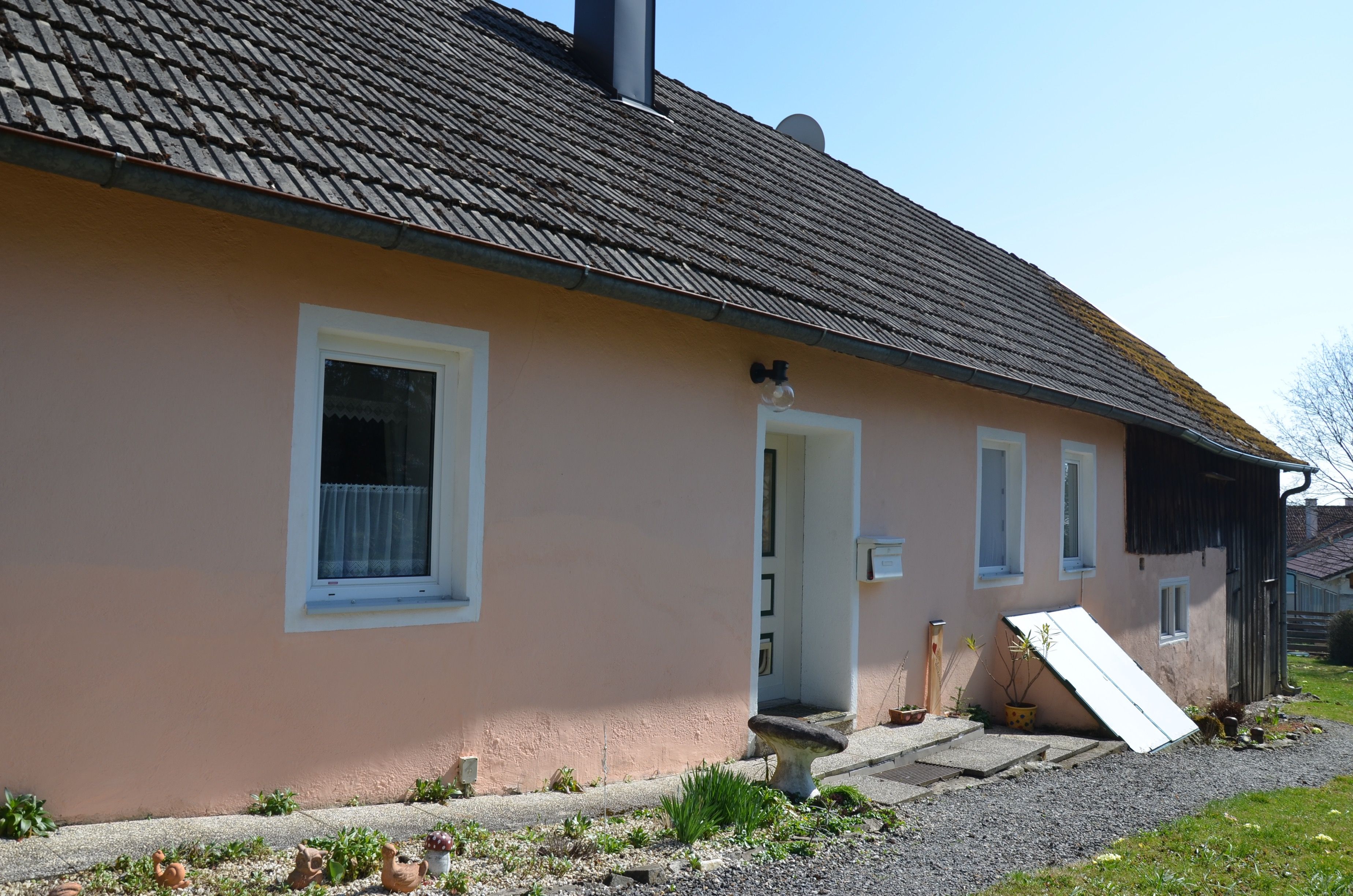 Ein einfaches, einstöckiges Haus mit rosa Fassade und grauem Dach. Vor dem Haus sind Pflanzen und Dekorationen zu sehen.