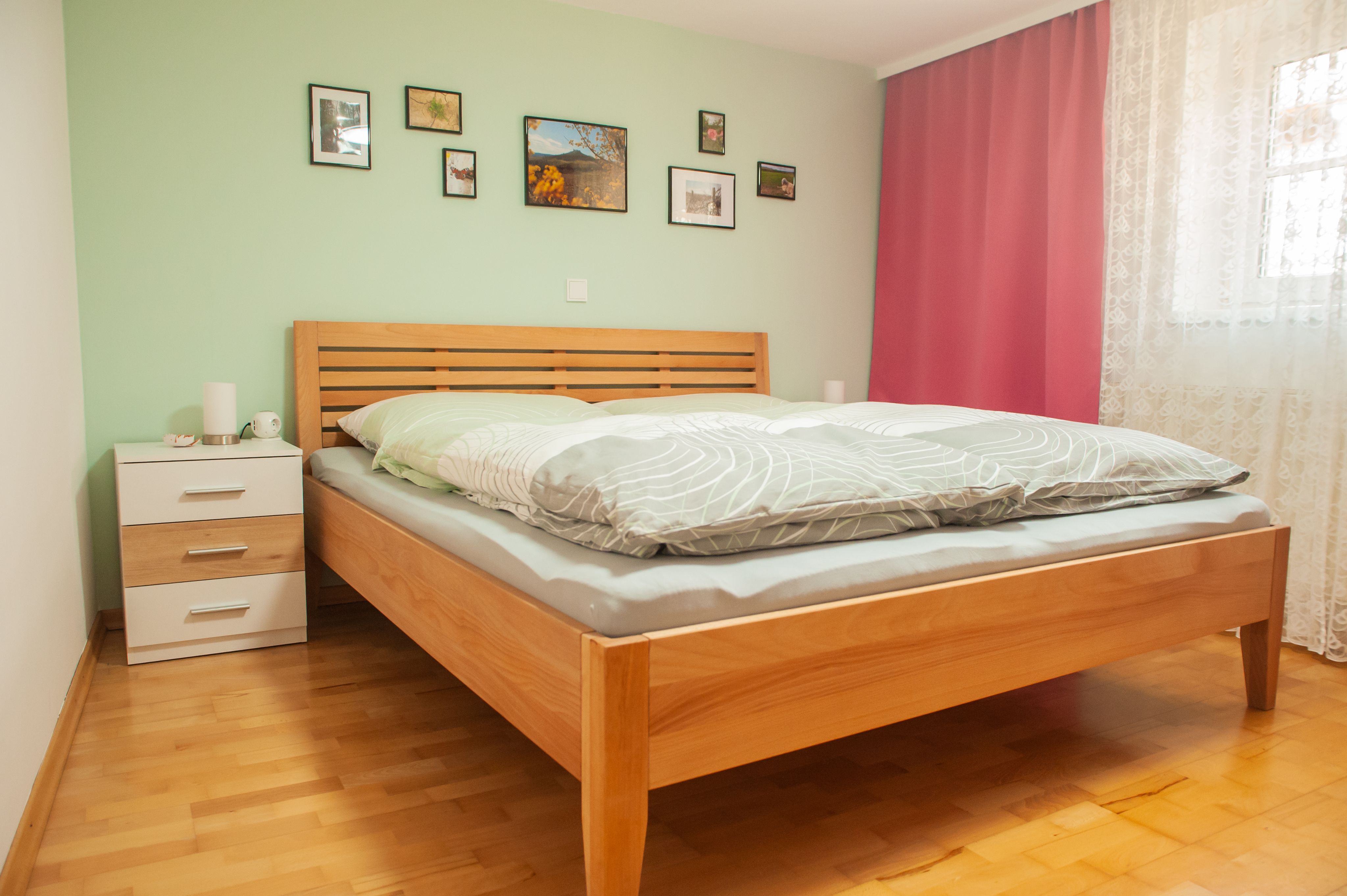 Ein Schlafzimmer mit einem Holzbett, grüner Wand, Bildern, Nachttisch und rosa Vorhängen.