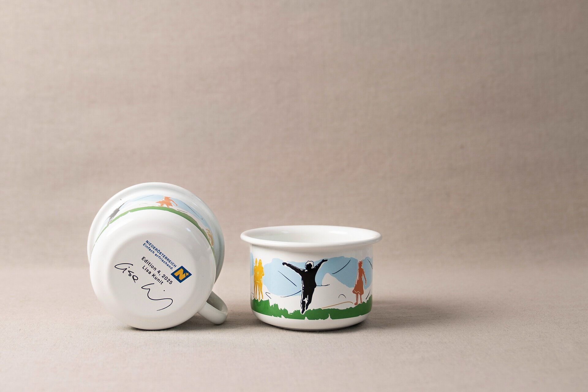 Künstlerisch gestaltet Tasse von Riess Keramik. Einmal in der Frontalansicht mit buntem Motiv inspiriert aus Niederösterreich. Zweite Tasse liegt daneben auf der Seite und zeigt den Boden mit Unterschrift der Künstlerin. 