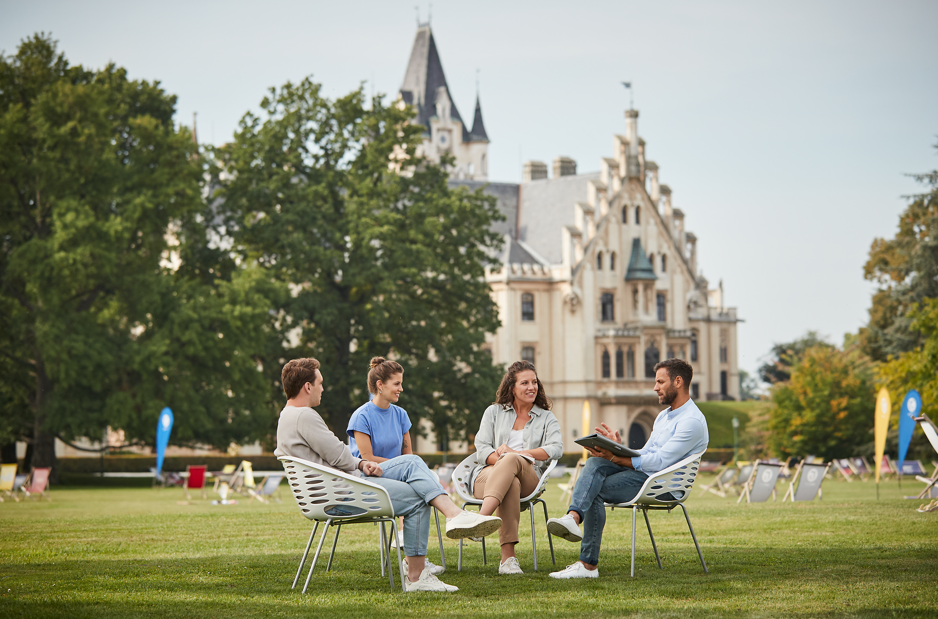 Inmitten einer malerischen Kulisse entspannen sich vier Freunde auf modernen Stühlen, umgeben von üppigem Grün und der majestätischen Architektur im Hintergrund. Die sanfte Brise und das Lachen der Gruppe schaffen eine einladende Atmosphäre, die den perfekten Rahmen für unvergessliche Momente bietet.