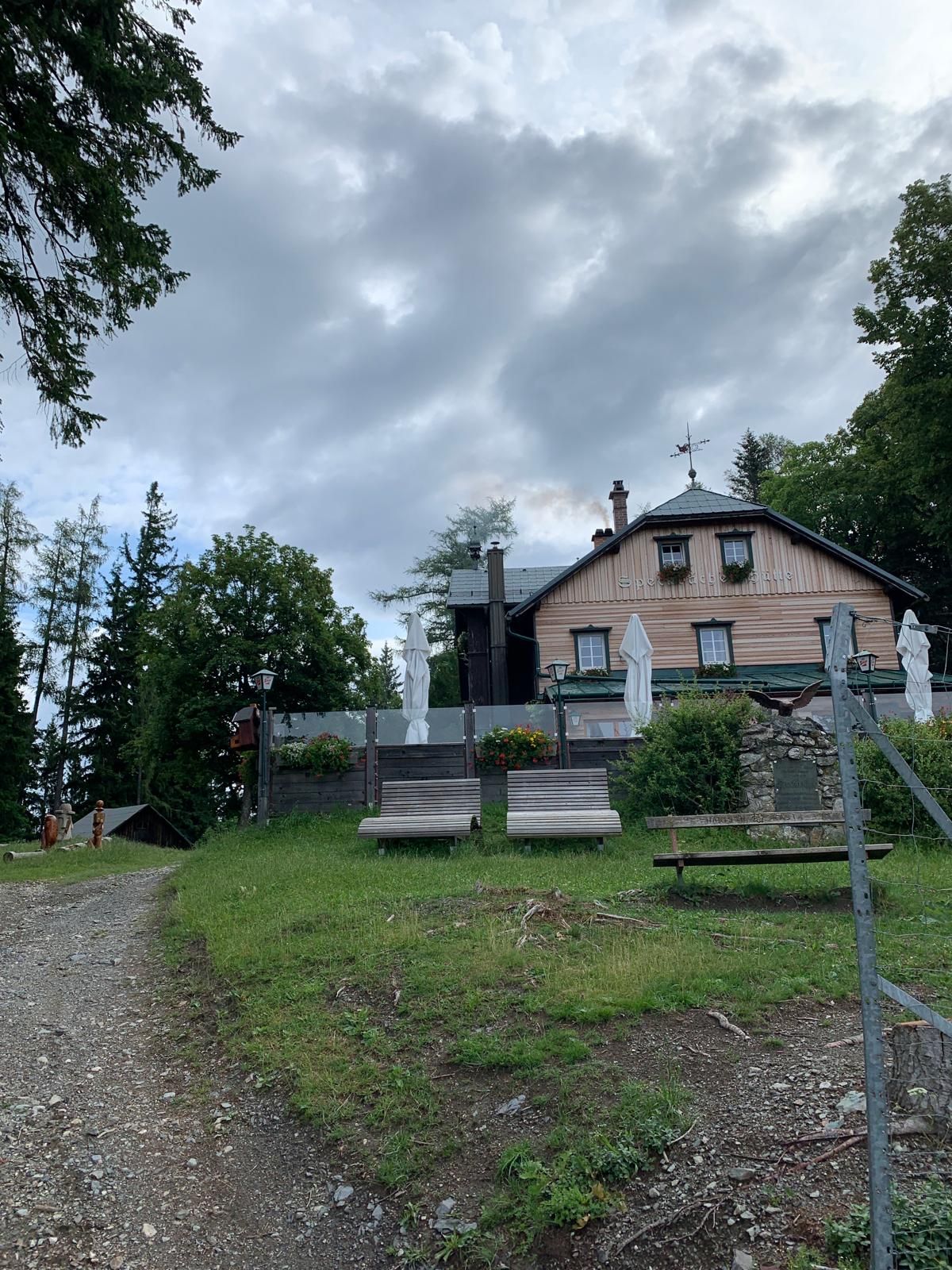Eine Berghütte mit Terrasse und Liegestühlen auf einer Wiese, umgeben von Bäumen und bewölktem Himmel.
