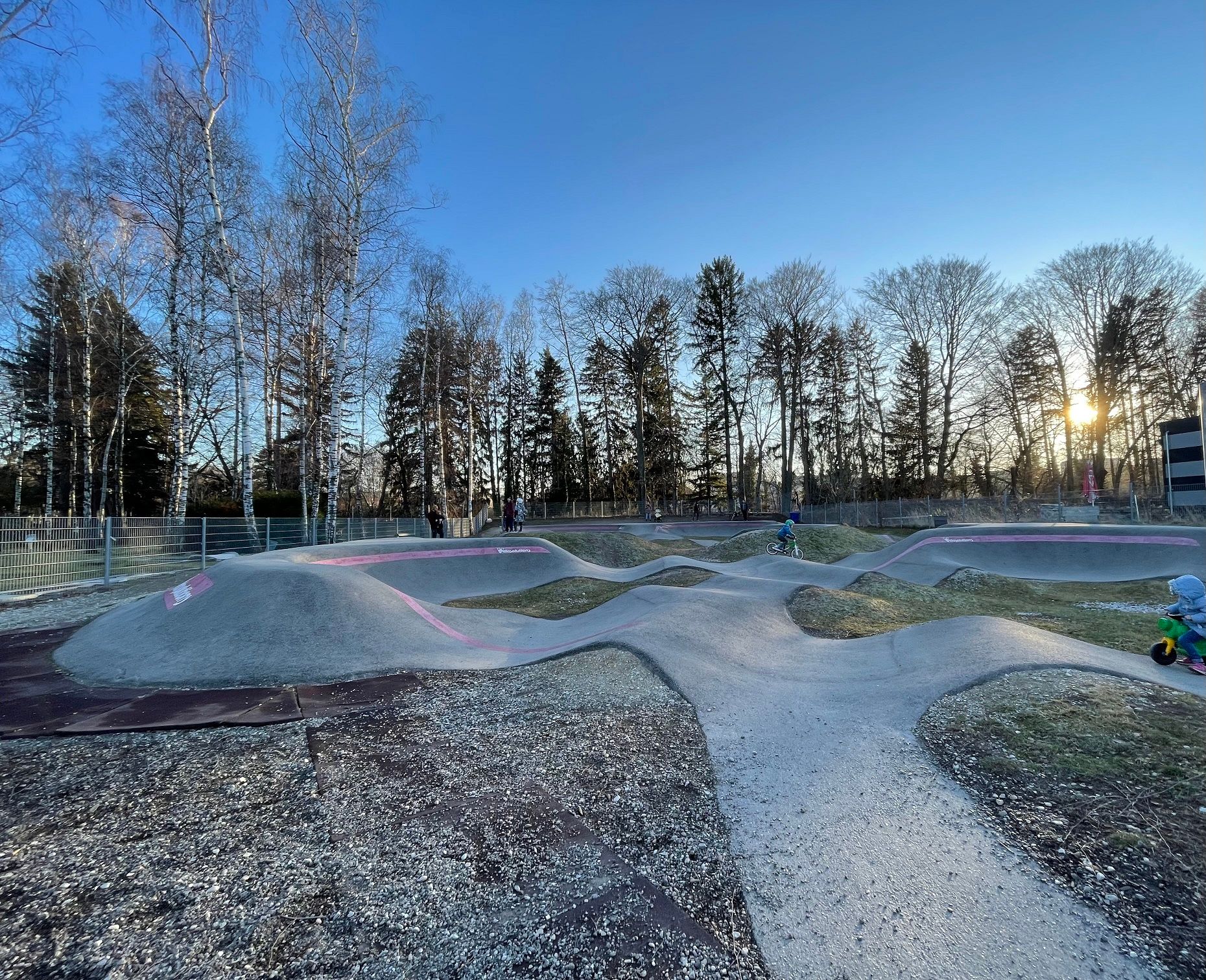 Pumptrack in Neunkirchen mit Kindern auf Fahrrädern und Laufrädern, umgeben von Bäumen bei Sonnenuntergang.