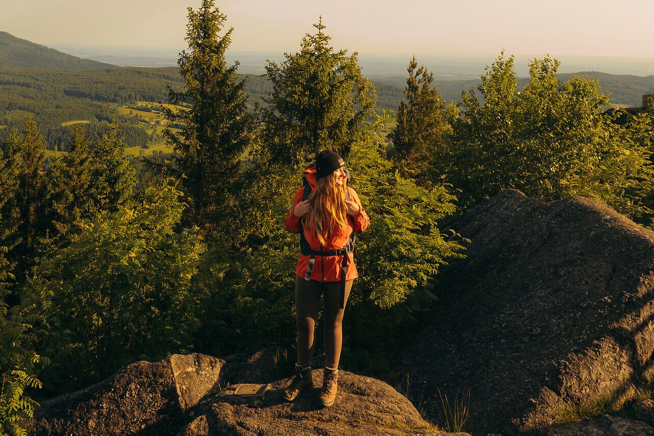 Ein Wanderer steht auf einem Felsen mit Ausblick auf bewaldete Hügel, aufgenommen im Waldviertel, gekrönt von einem klaren Himmel und dem Licht der aufgehenden Sonne.