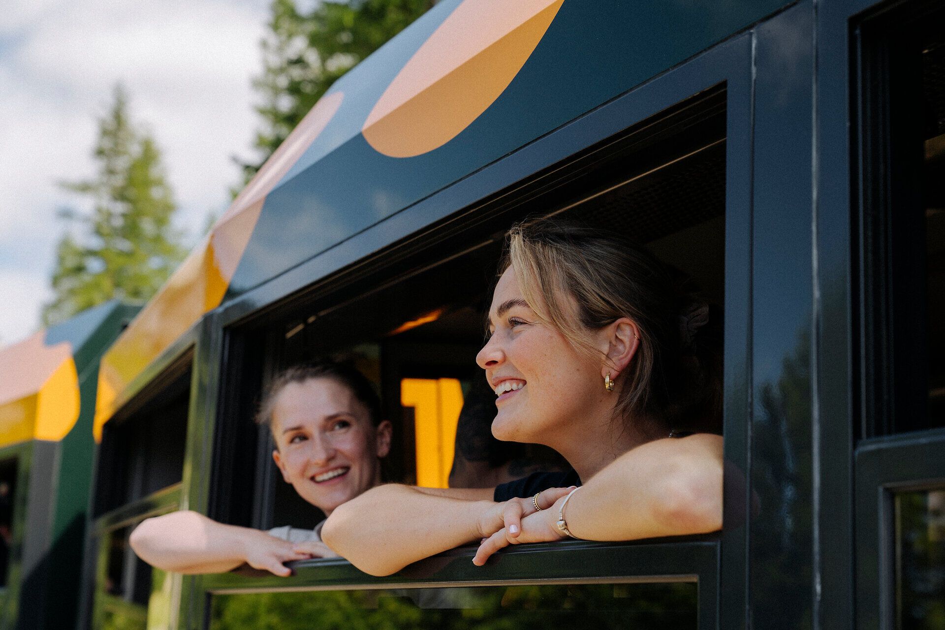 Zwei fröhliche Frauen schauen aus dem Fenster des Salamander-Zuges der Schneebergbahn, während die Landschaft aus Bäumen im Hintergrund vorbeizieht, was Freude und Neugier auf das bevorstehende Reiseerlebnis widerspiegelt.