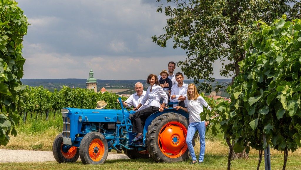 Eine Gruppe von Menschen auf einem blauen Traktor in einem Weinberg.