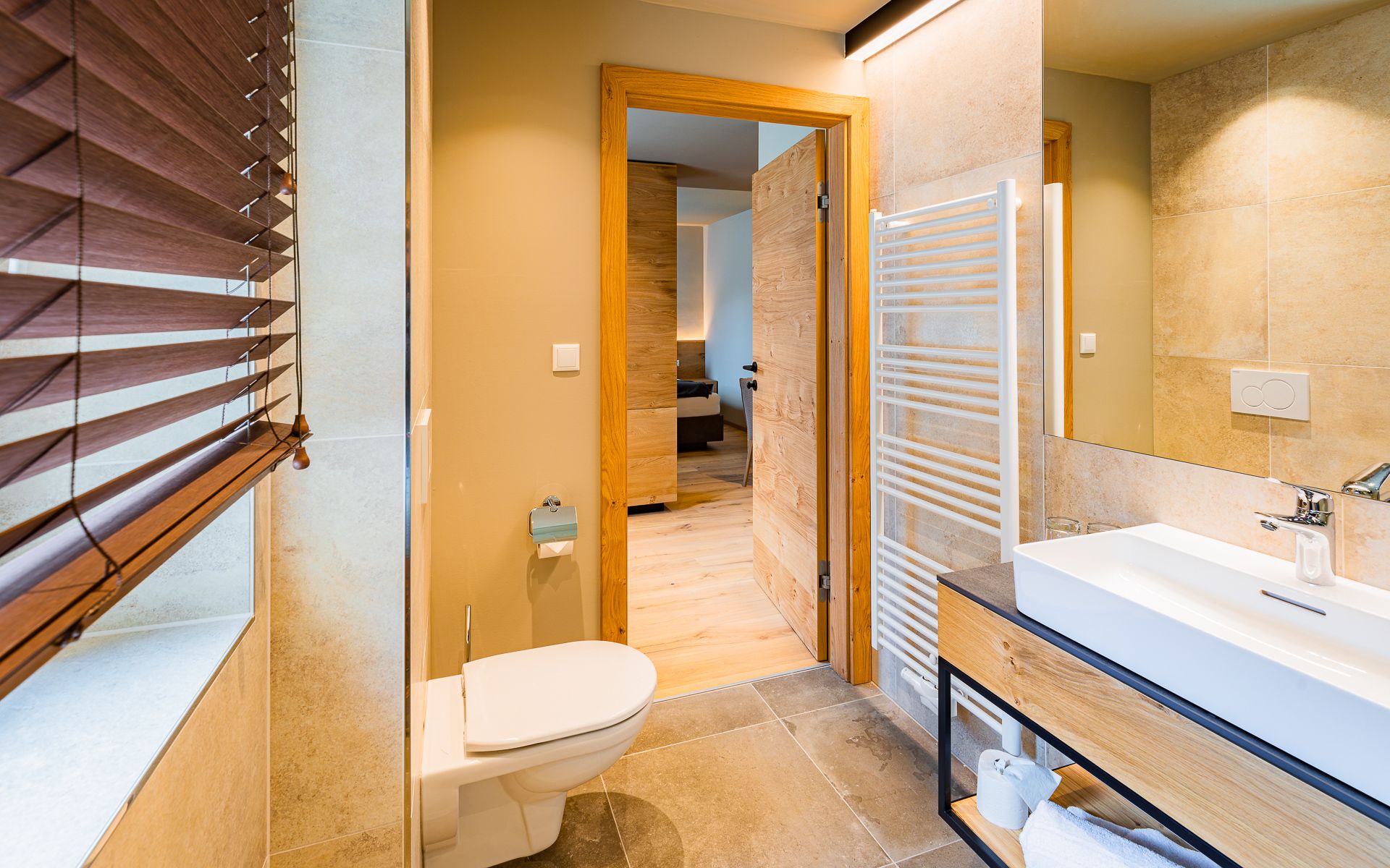 Modernes Badezimmer mit Toilette, Waschtisch, Spiegel und Holzdetails; durch die offene Tür ist ein Schlafzimmer zu sehen.