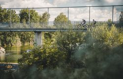 Zwei Personen mit Helmen fahren mit Fahrrädern über eine Brücke über die Traisen; rundherum sind Bäume und Sträucher zu sehen.