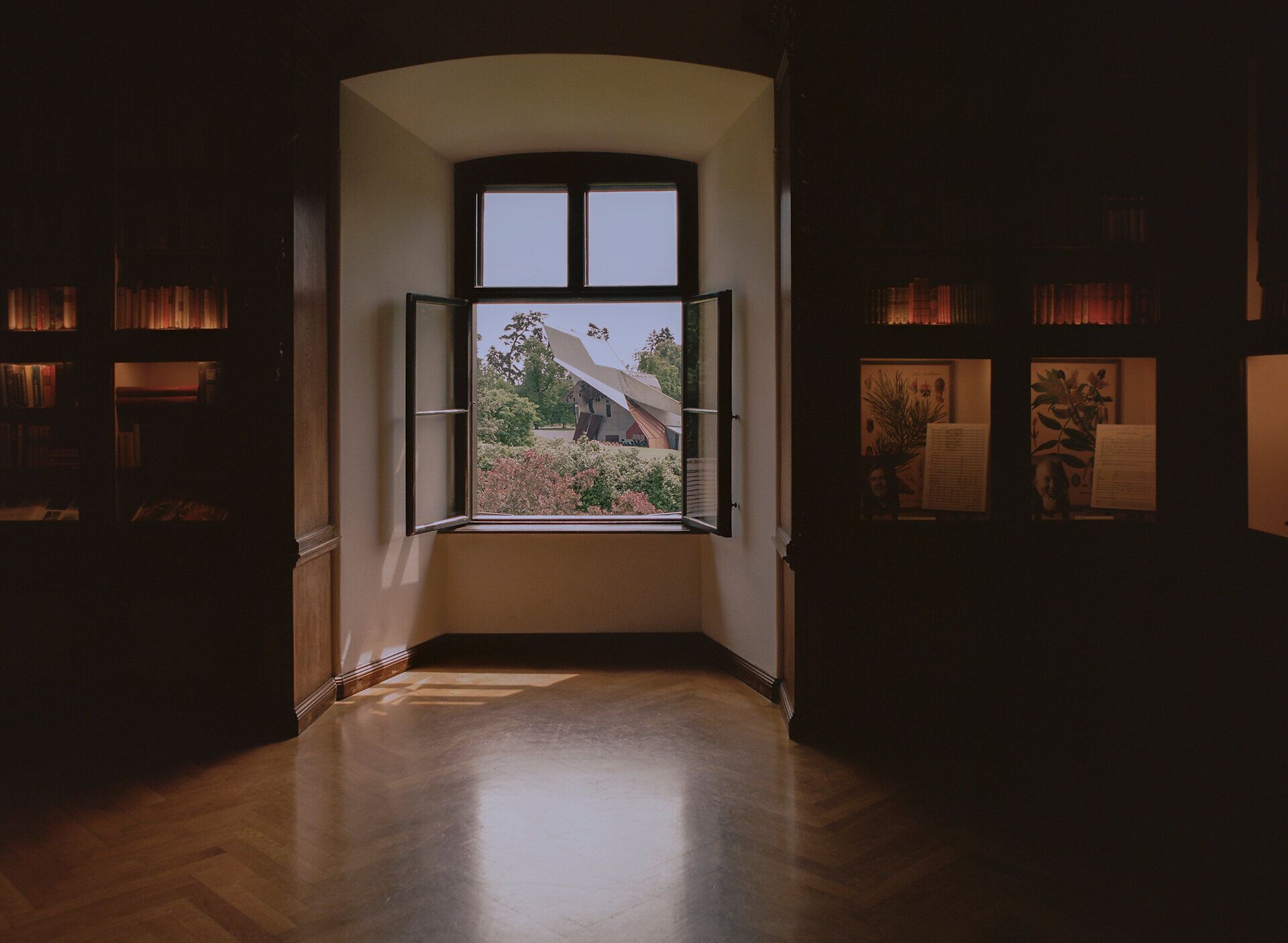 Blick aus einem historischen Raum im Schloss Grafenegg durch ein geöffnetes Fenster auf den modernen Wolkenturm, mit Bücherregalen und beleuchteten Vitrinen im Vordergrund.