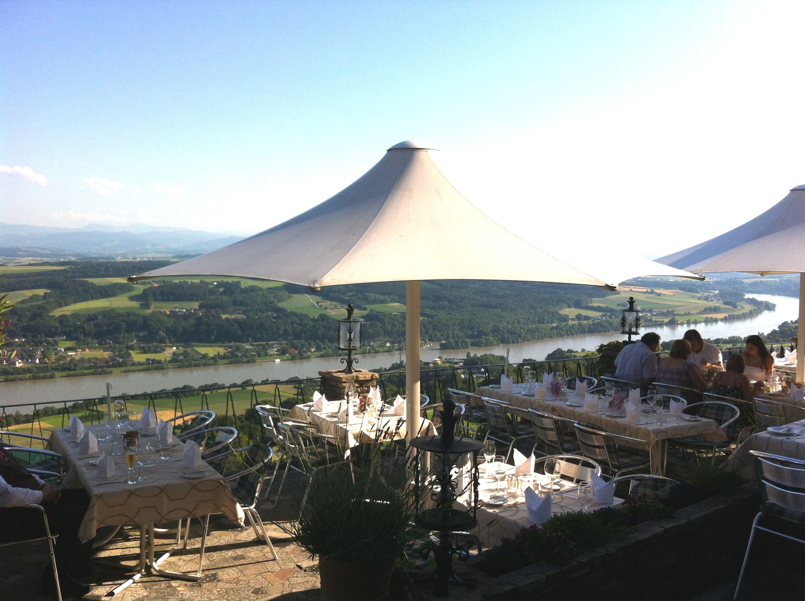 Terrasse mit Tischen und Sonnenschirmen, Blick auf Fluss und Landschaft.