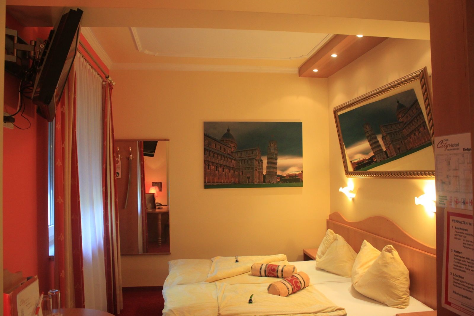 Hotelzimmer mit Doppelbett, Bildern des Schiefen Turms von Pisa und einem Fernseher an der Wand.