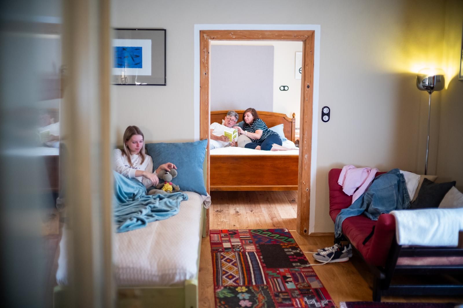 Innenansicht einer Ferienwohnung mit zwei Zimmern. Im Vordergrund sitzt ein Mädchen auf einem Bett, im Hintergrund lesen zwei Personen auf einem anderen Bett.