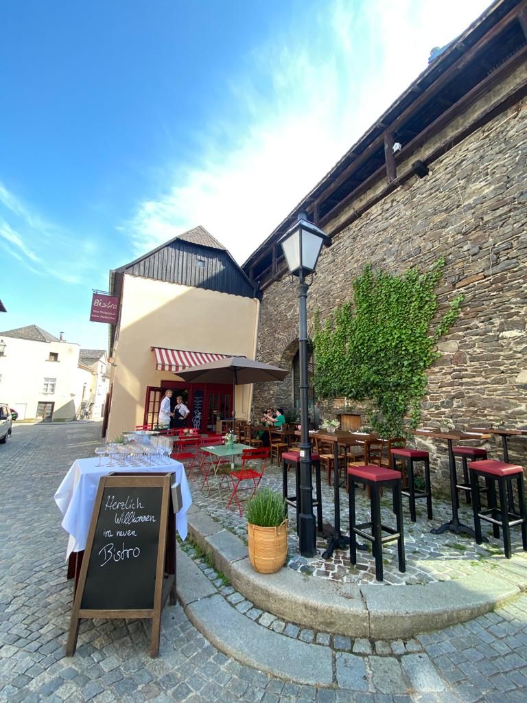 Ein gemütliches Bistro mit roten Stühlen und Tischen im Freien, umgeben von alten Steinmauern und Pflanzen.