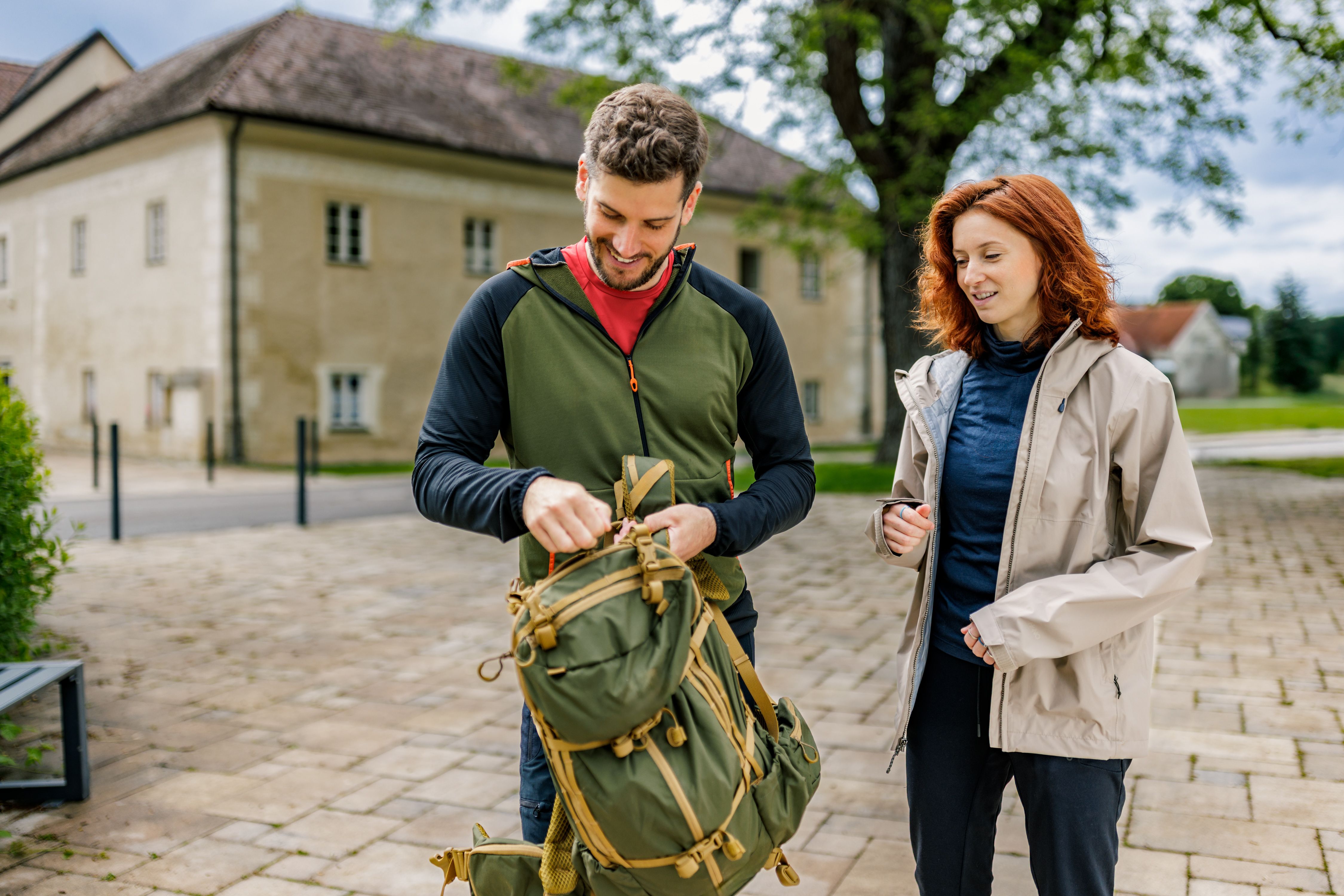 Ein Mann und eine Frau stehen draußen vor dem Schloss Katzelsdorf. Der Mann packt einen grünen Rucksack, während die Frau zuschaut. Beide tragen Outdoor-Kleidung.