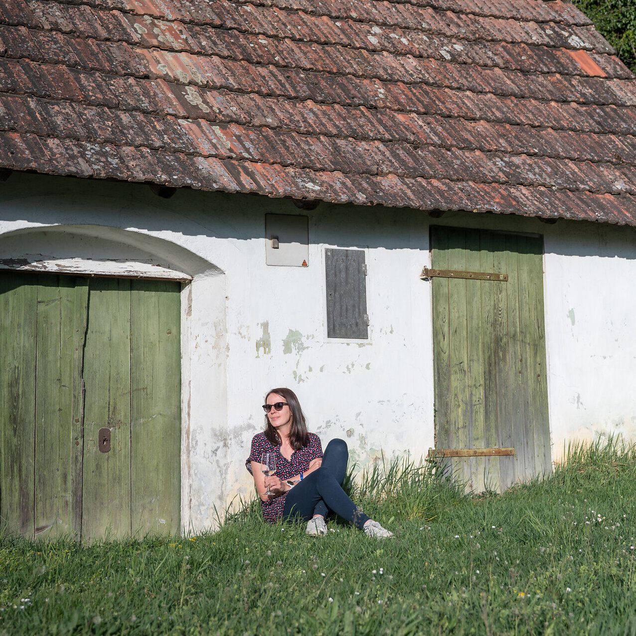 Eine Frau sitzt angelehnt an ein Presshaus in einer Wiese und genießt ein Glas Weißwein. Ihr Blick ist zu den frühlingshaften Sonnenstrahlen gereichtet.