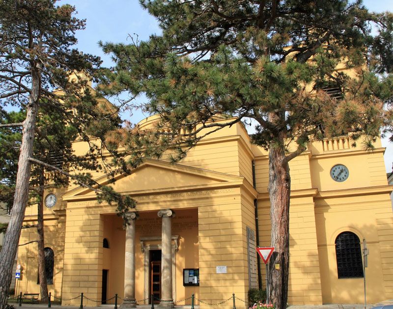 Außenansicht der Pfarre Hinterbrühl mit gelber Fassade und Uhr.