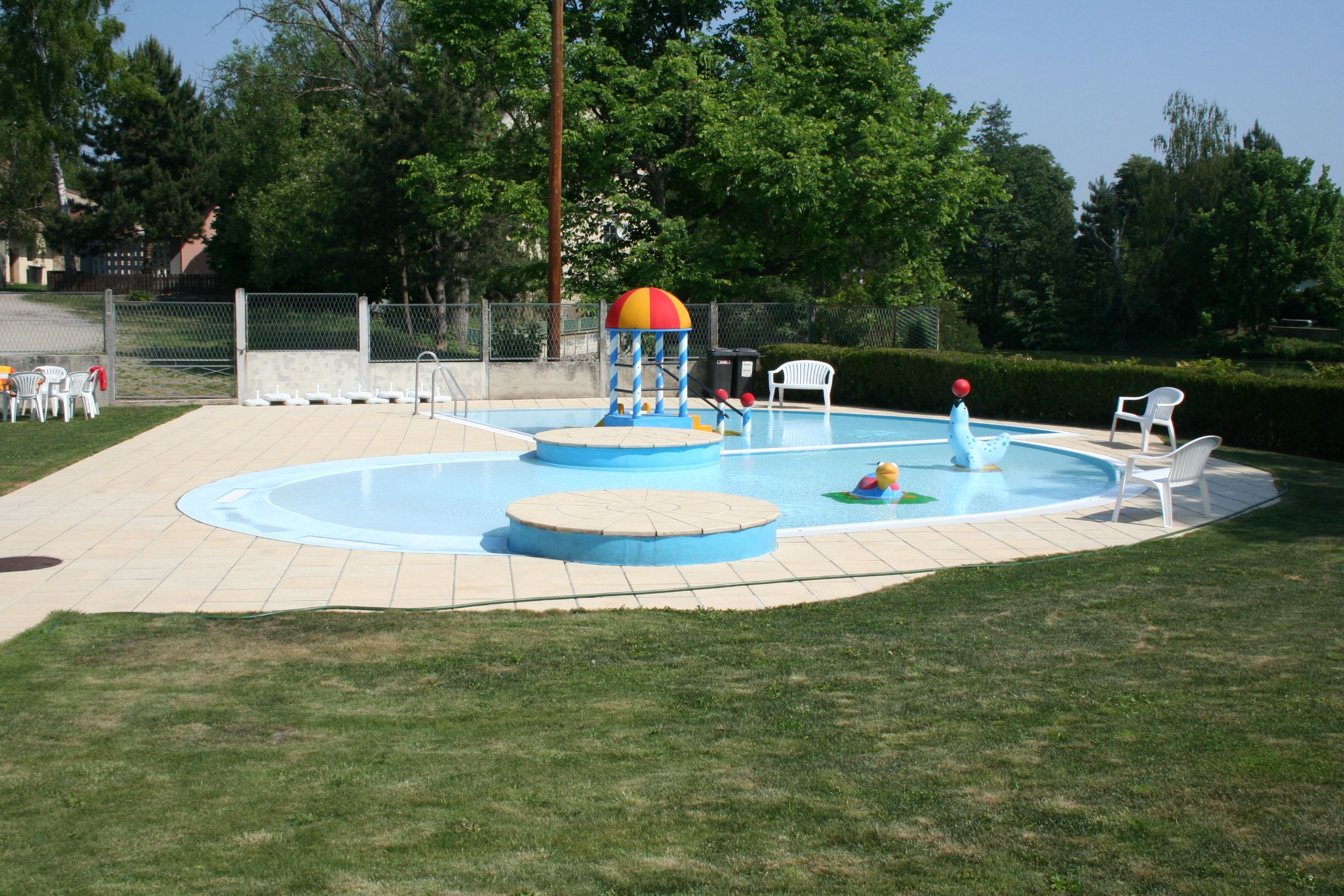 Ein Kinderplanschbecken mit bunten Wasserspielzeugen und weißen Plastikstühlen auf einer Wiese.