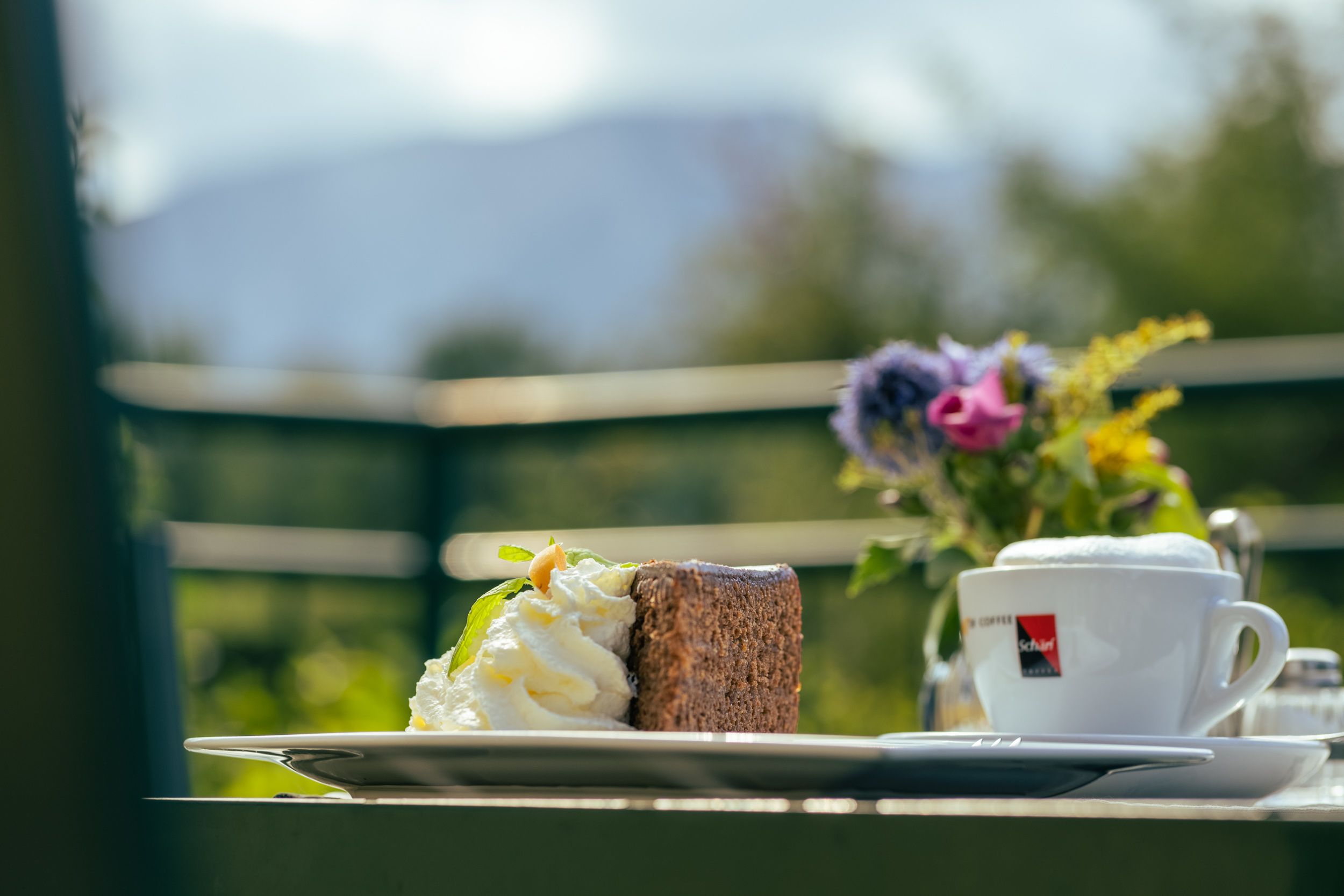 Ein Stück Kuchen mit Sahne und eine Tasse Kaffee auf einem Tisch im Freien, im Hintergrund unscharfe Berge.
