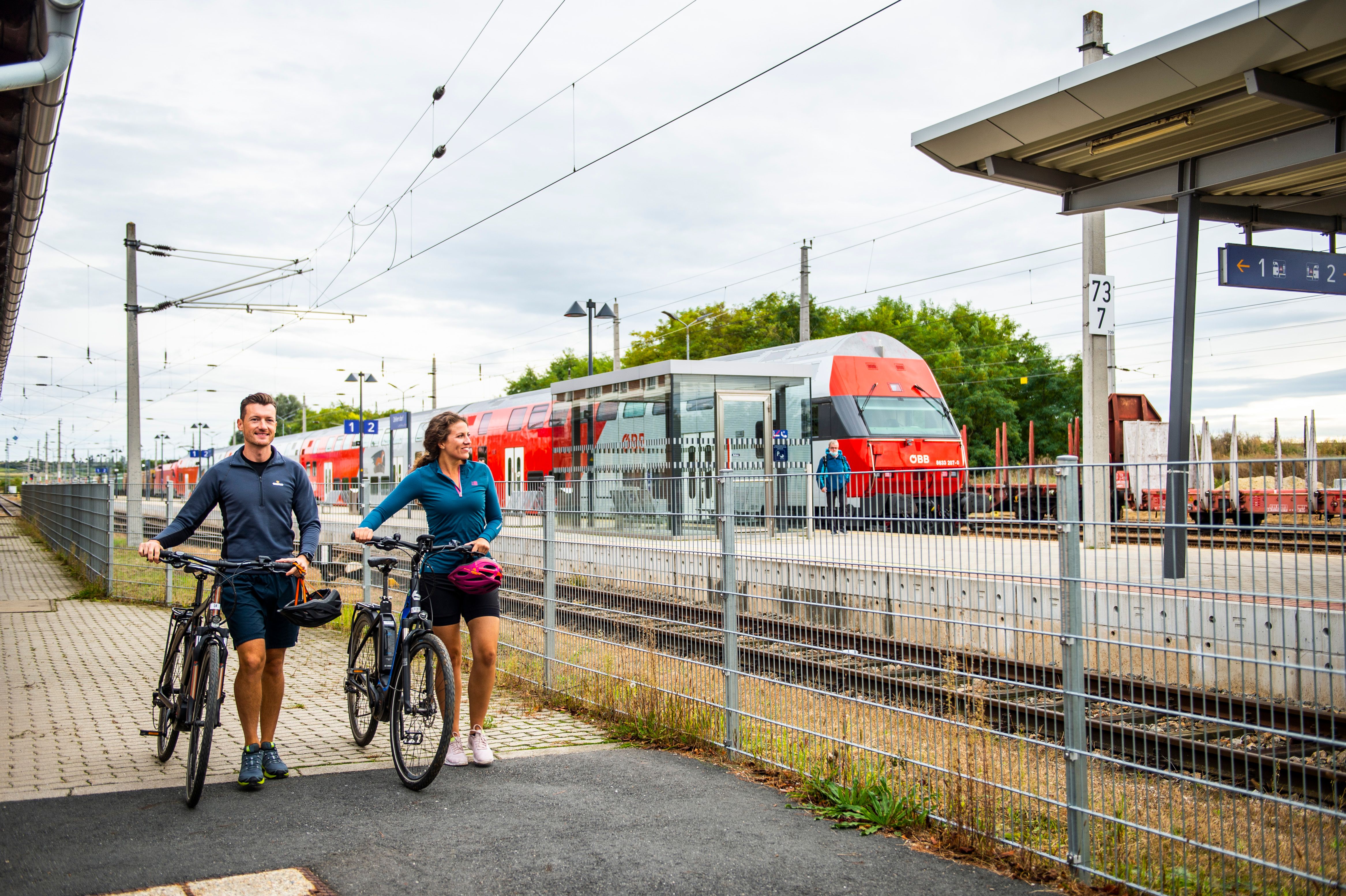 Zwei Personen mit Fahrrädern stehen auf einem Bahnsteig neben einem ÖBB-Zug.