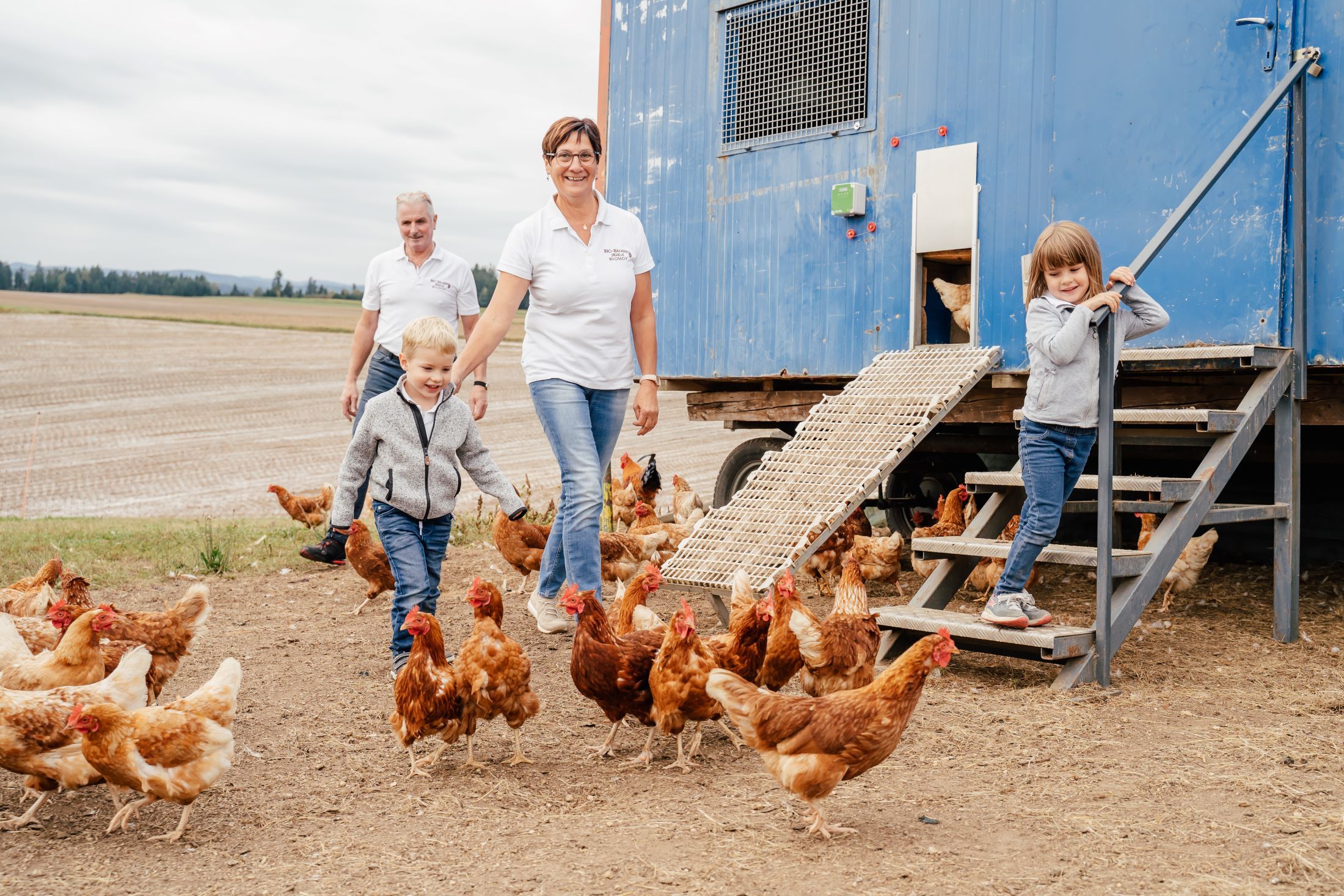 Familie mit Hühnern vor einem blauen Hühnerstall auf einem Feld.