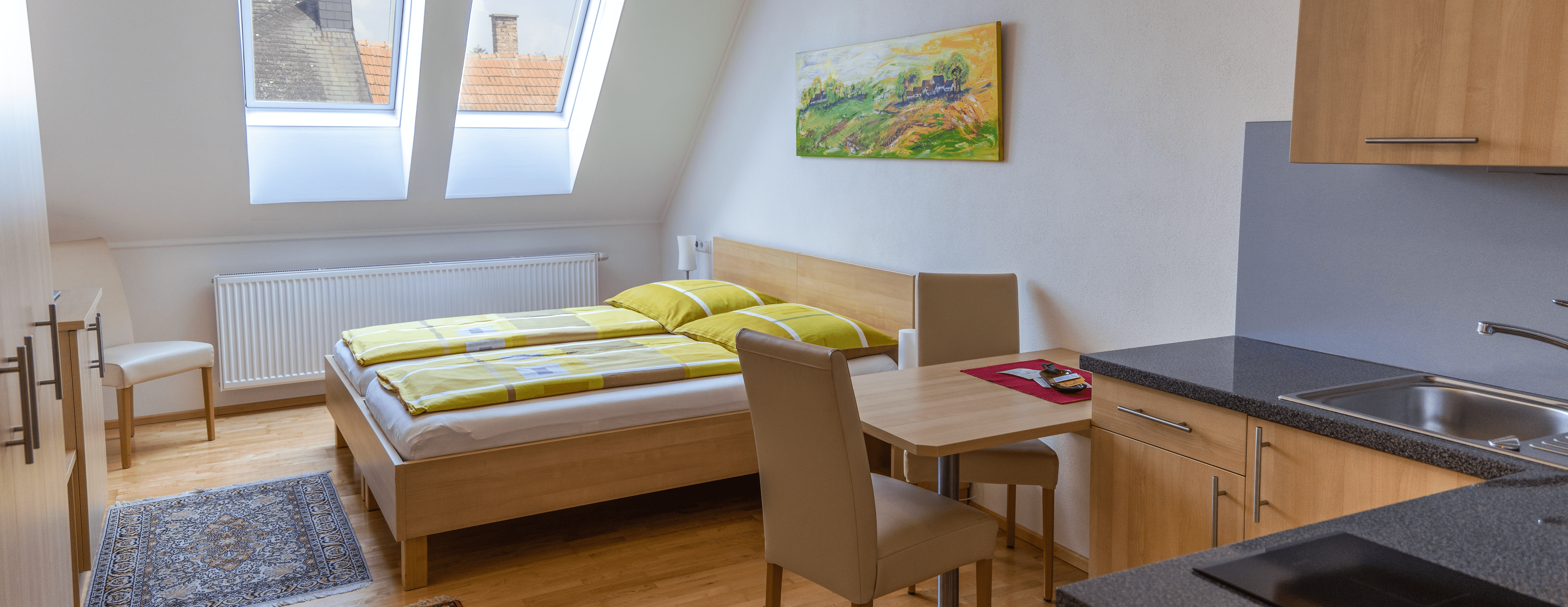 Doppelzimmer mit Küchenzeile, Bett, Tisch und Stühlen.