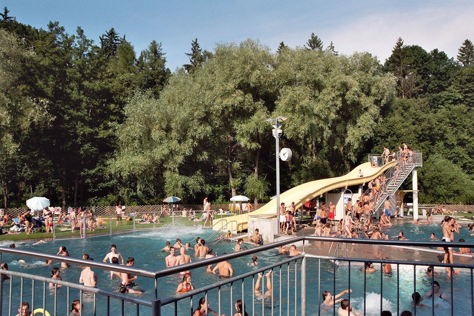 Freibad mit vielen Menschen, Rutsche und Bäumen im Hintergrund.