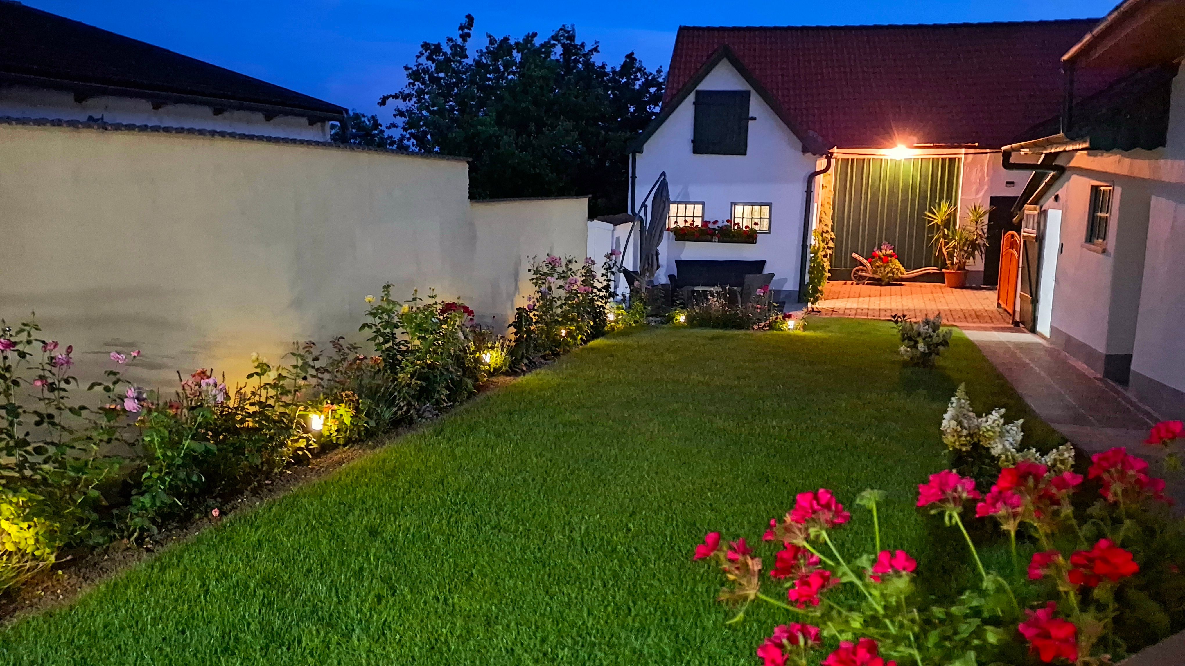 Ein beleuchteter Innenhof bei Nacht mit gepflegtem Rasen, Blumenbeeten und einem kleinen Gebäude im Hintergrund.