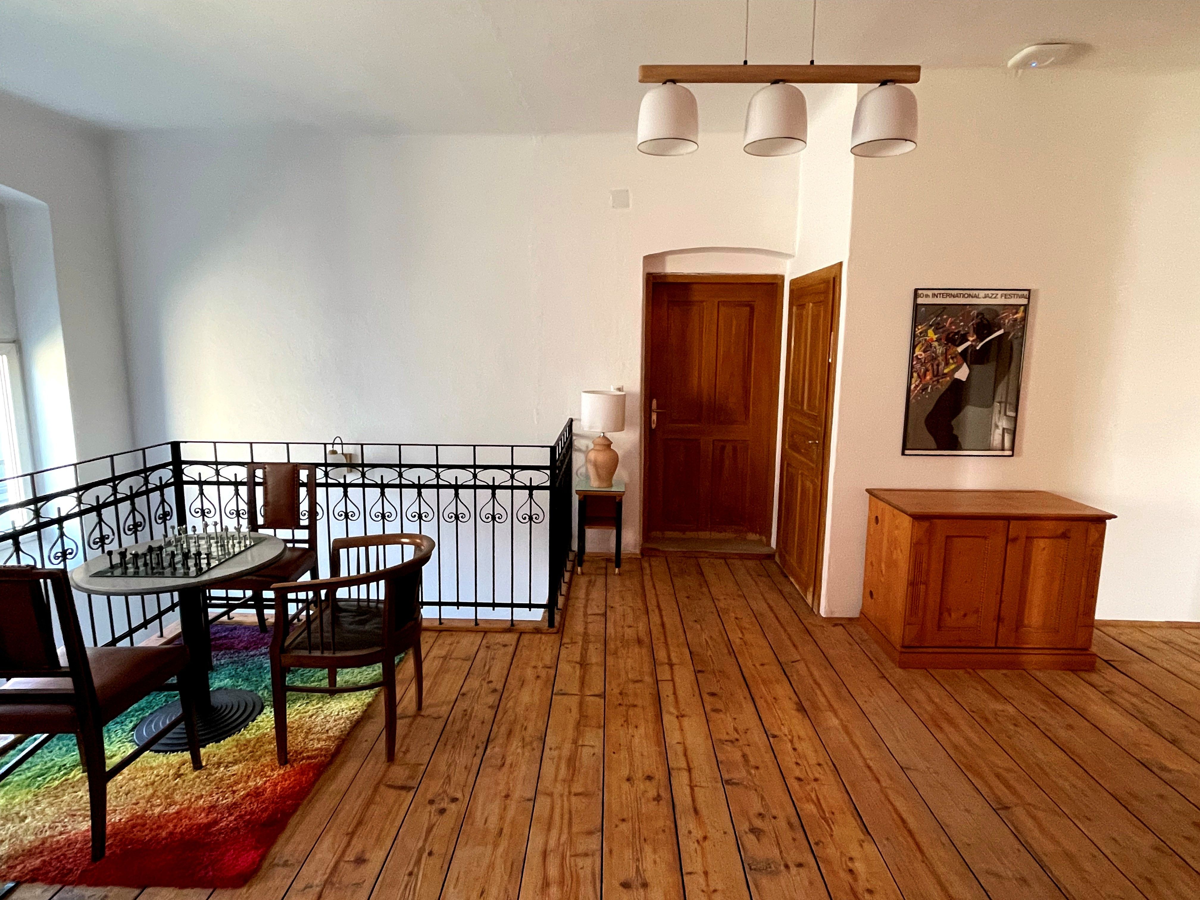 Ein gemütlicher Vorraum mit Holzboden, einem Tisch mit Schachbrett, zwei Stühlen und einem bunten Teppich. An der Wand hängt ein Jazz-Festival-Poster.