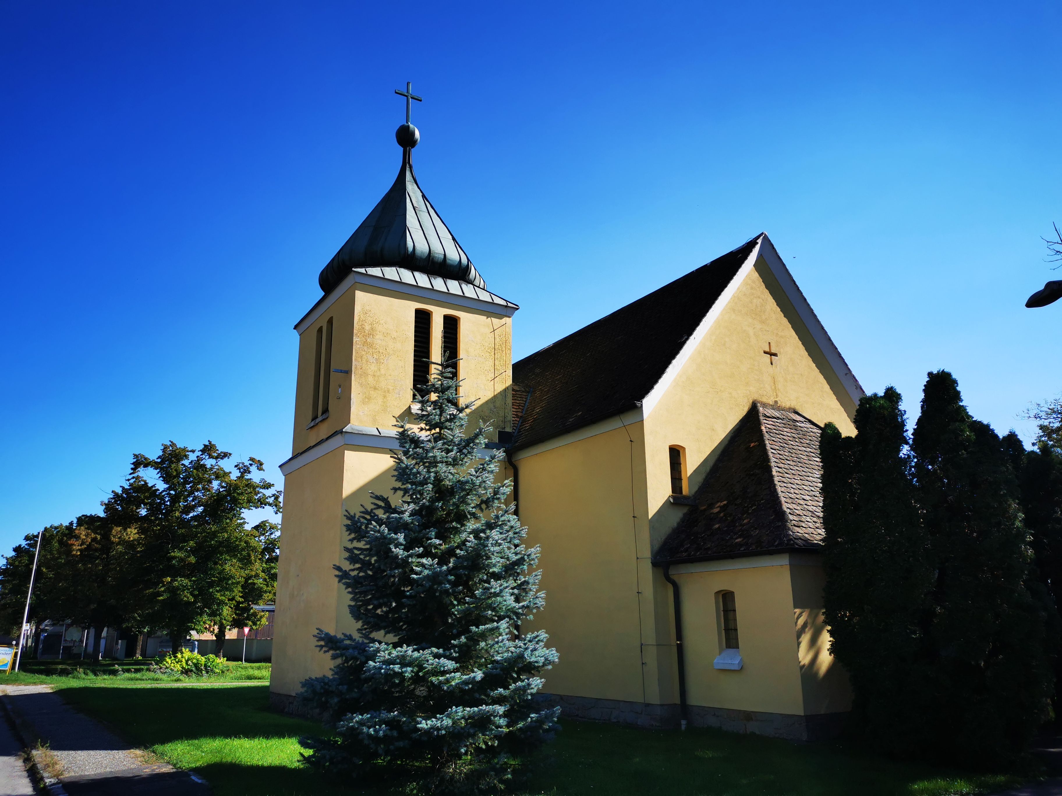 Gelbe Kapelle mit Turm und Kreuz, umgeben von Bäumen, unter blauem Himmel.