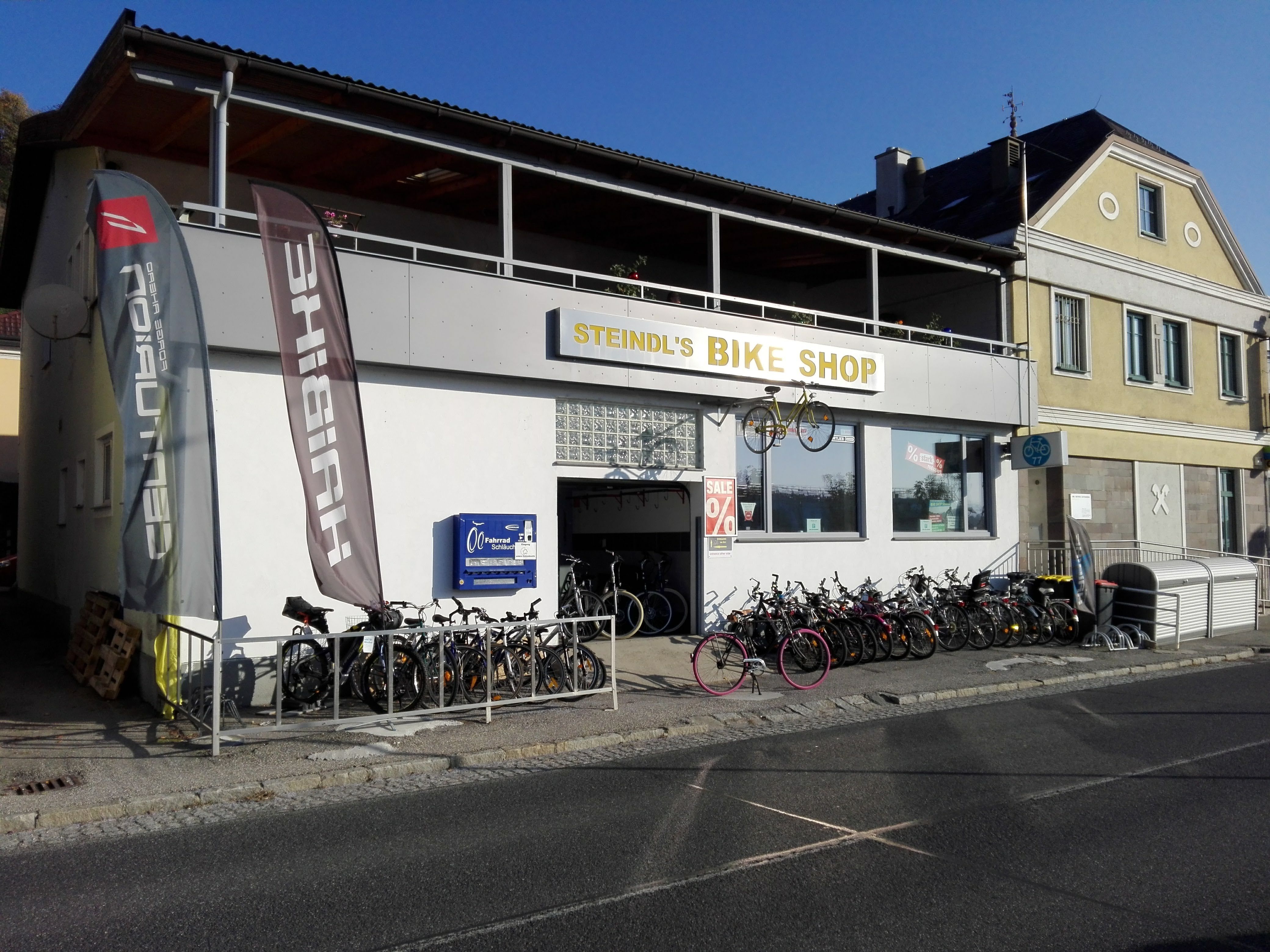 Fahrradgeschäft mit Fahrrädern vor dem Gebäude und Werbefahnen.
