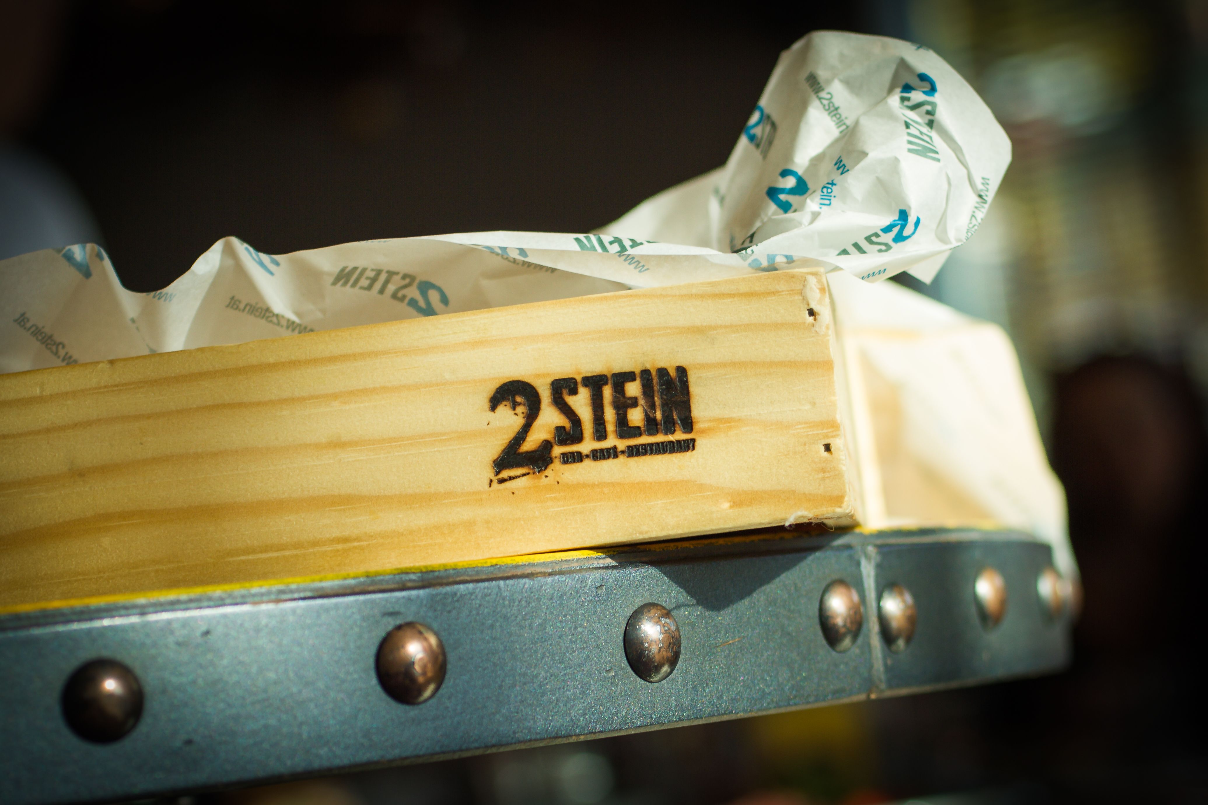 Holzkiste mit 2Stein-Logo und Papier im Hintergrund.