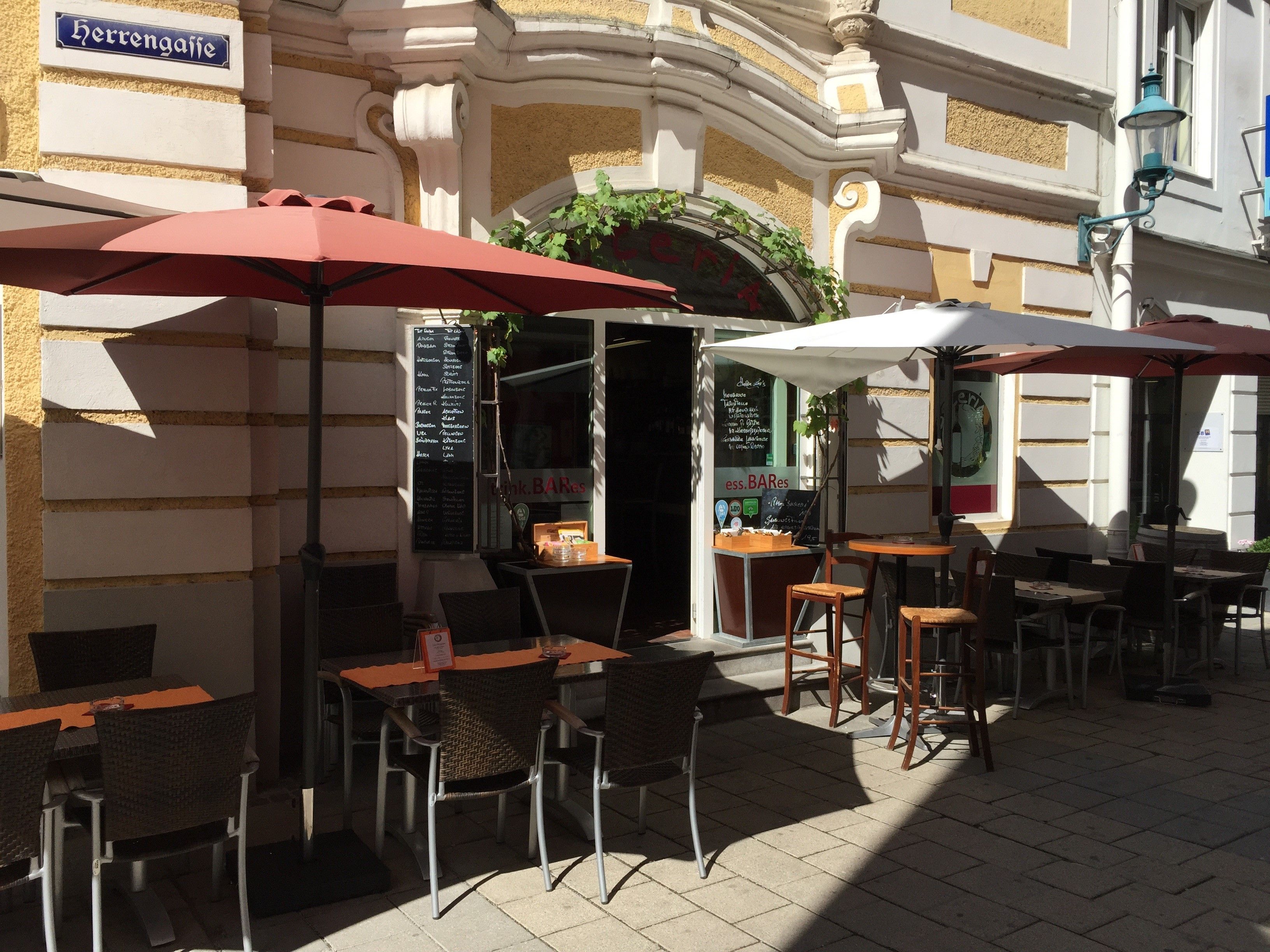 Außenbereich eines Restaurants mit Tischen, Stühlen und Sonnenschirmen in einer sonnigen Gasse.