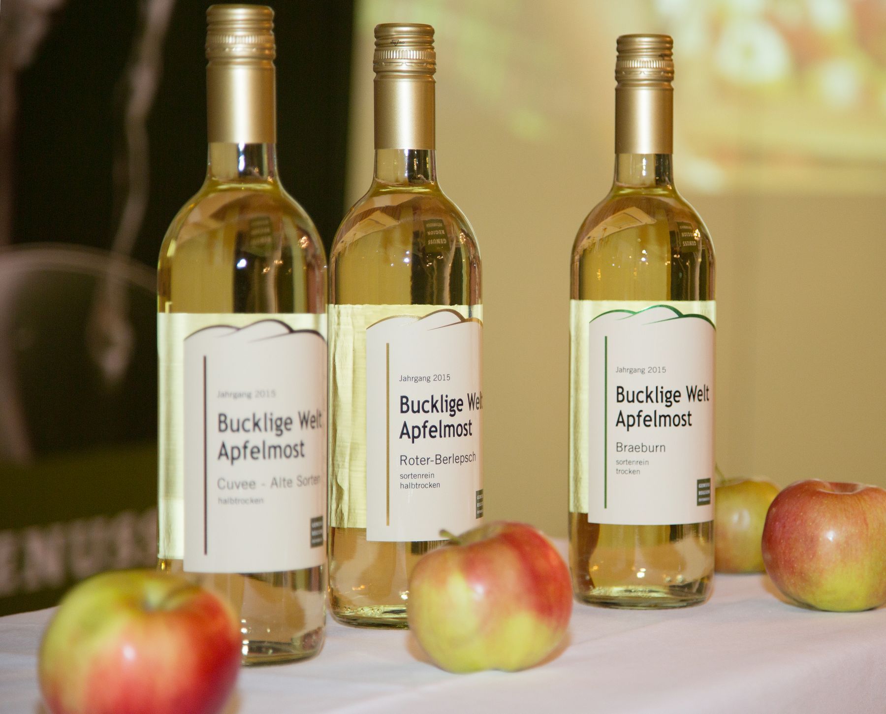 Drei Flaschen Bucklige Welt Apfelmost auf einem Tisch mit Äpfeln.
