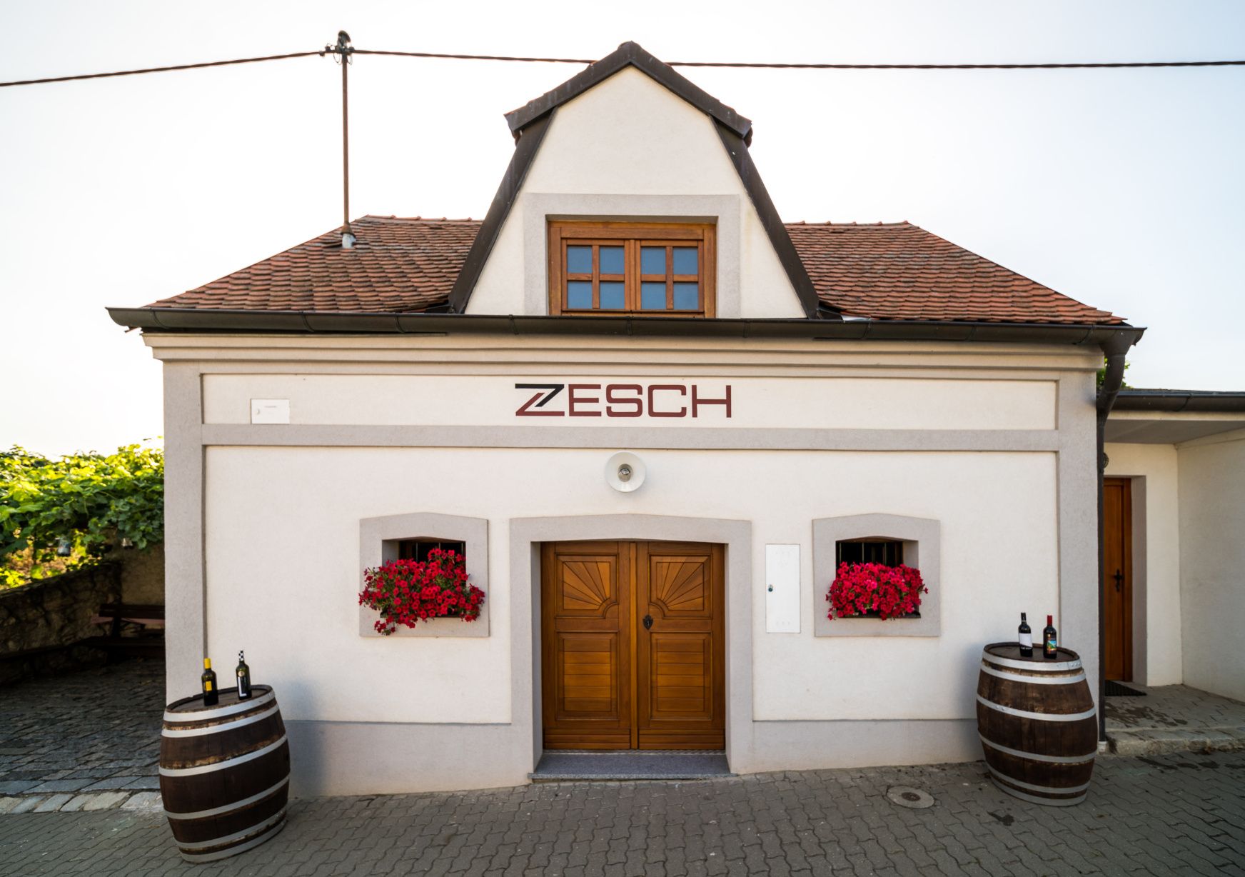 Kleines Gebäude mit der Aufschrift 'Zesch', zwei Weinfässern und roten Blumen.