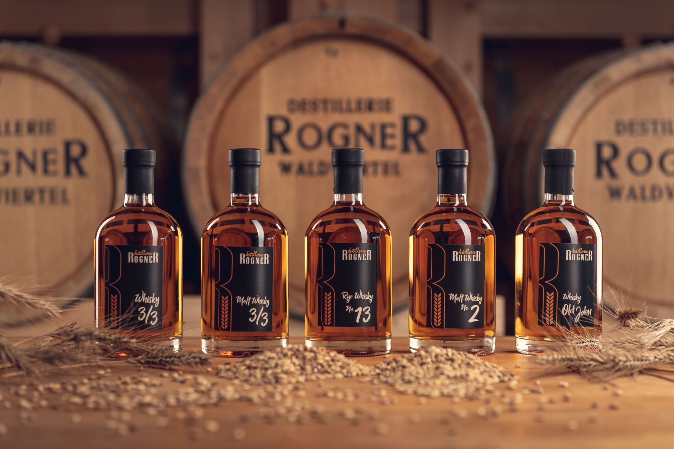 Fünf Whiskyflaschen der Destillerie Rogner vor Holzfässern.