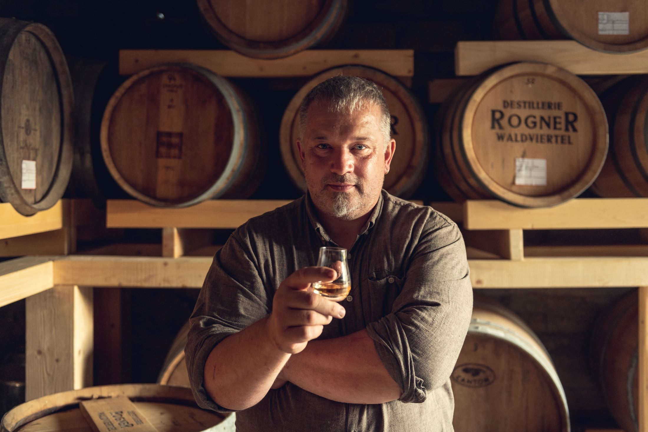 Mann in einer Destillerie mit einem Glas in der Hand vor Holzfässern.
