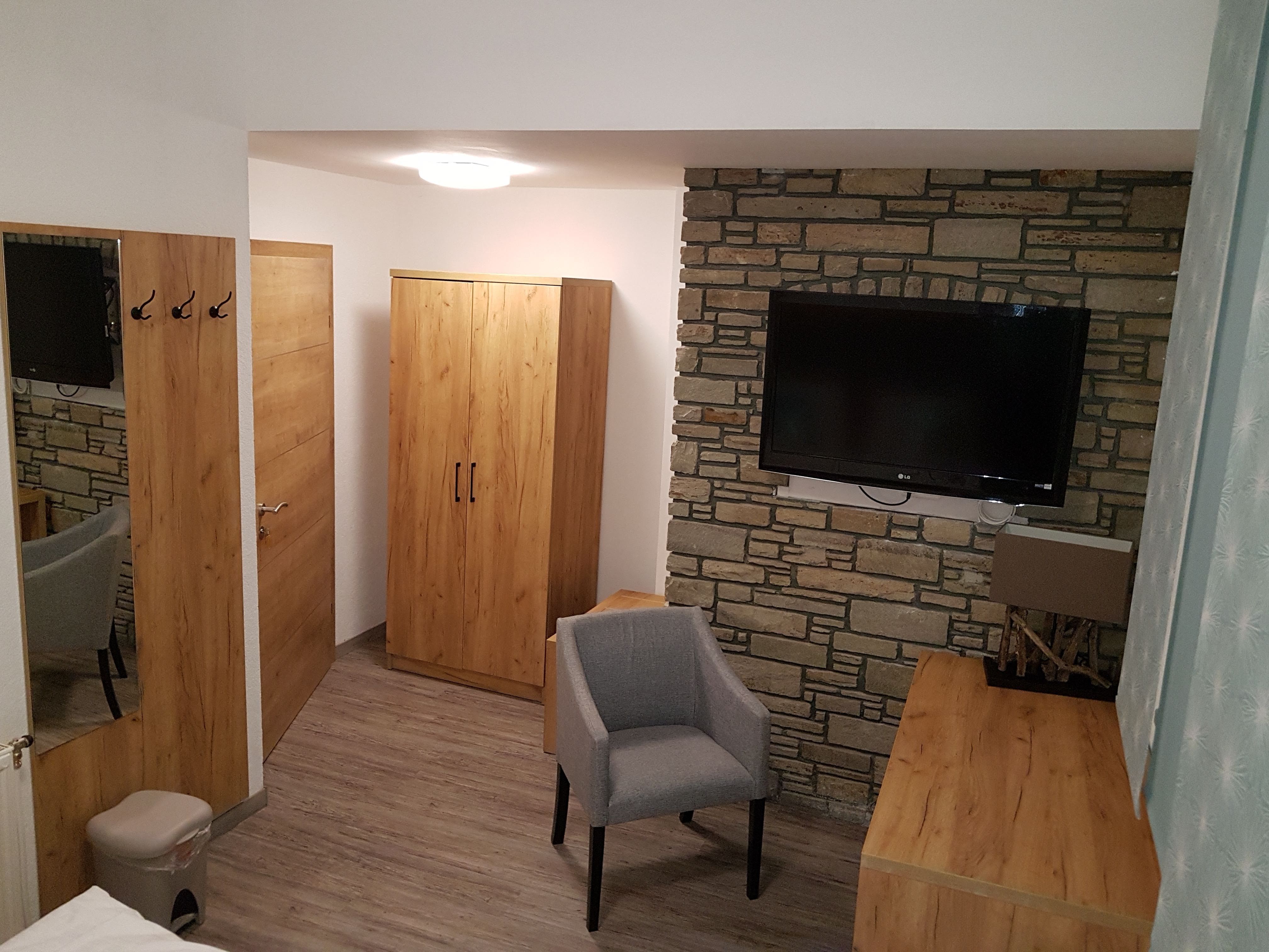 Hotelzimmer mit Holzmöbeln, grauem Sessel und Flachbildfernseher an einer Steinwand.