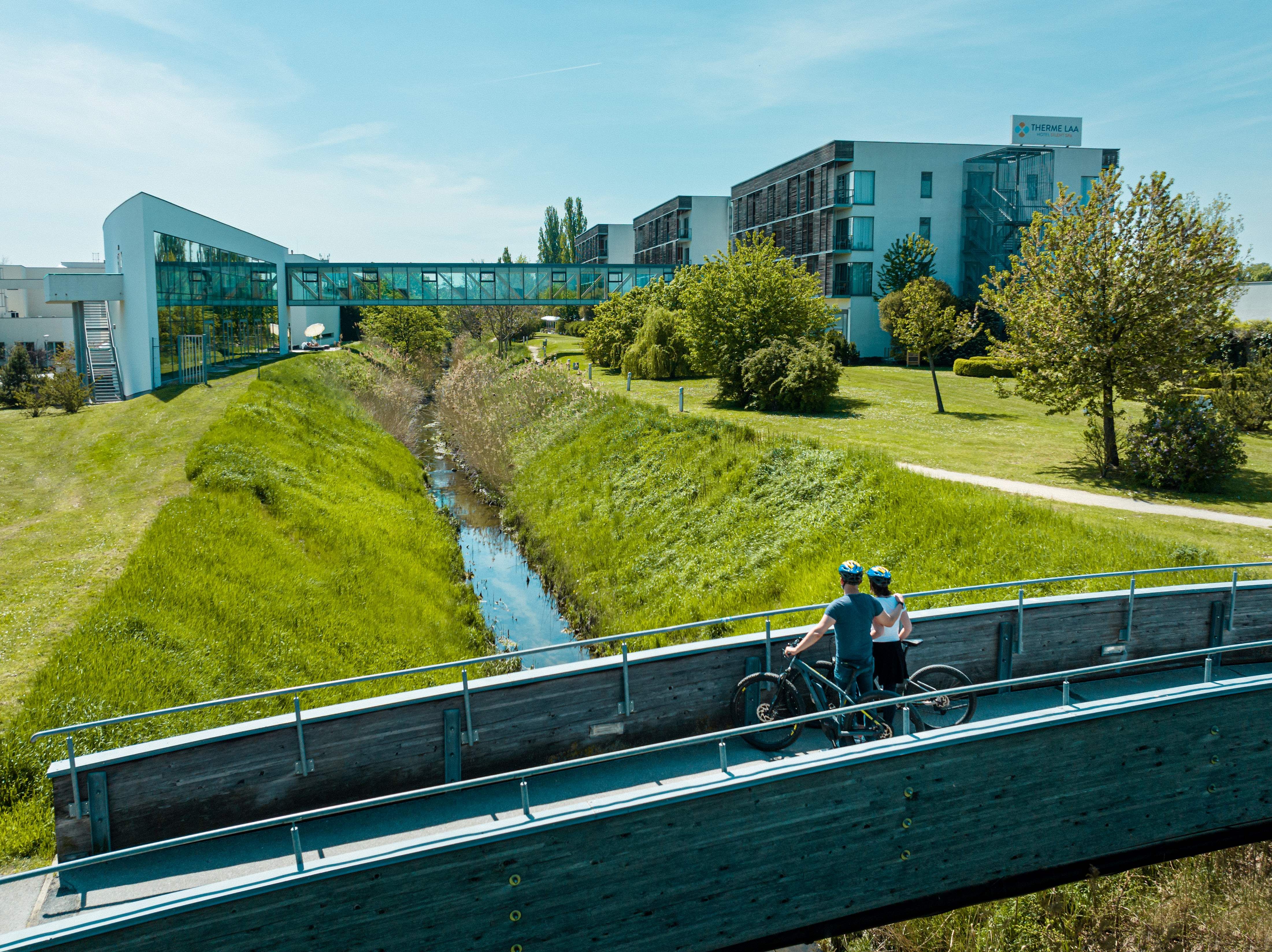 Zwei Radfahrer auf einer Brücke vor einem modernen Hotelgebäude mit Glasgang und grüner Umgebung.