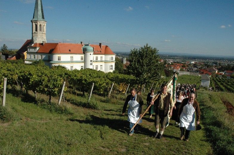 Menschen in traditioneller Kleidung gehen durch Weinberge in Gumpoldskirchen, im Hintergrund eine Kirche und Gebäude.
