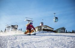 Zwei Wanderer gehen auf einem Weg bergauf, vorbei an einem Aussichtsturm und einem Gebäude, umgeben von Bäumen und einem klaren Himmel.