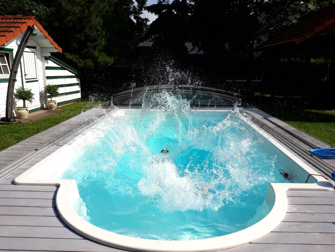 Ein Pool im Garten mit spritzendem Wasser, umgeben von Bäumen und einem kleinen Gebäude mit rotem Dach.