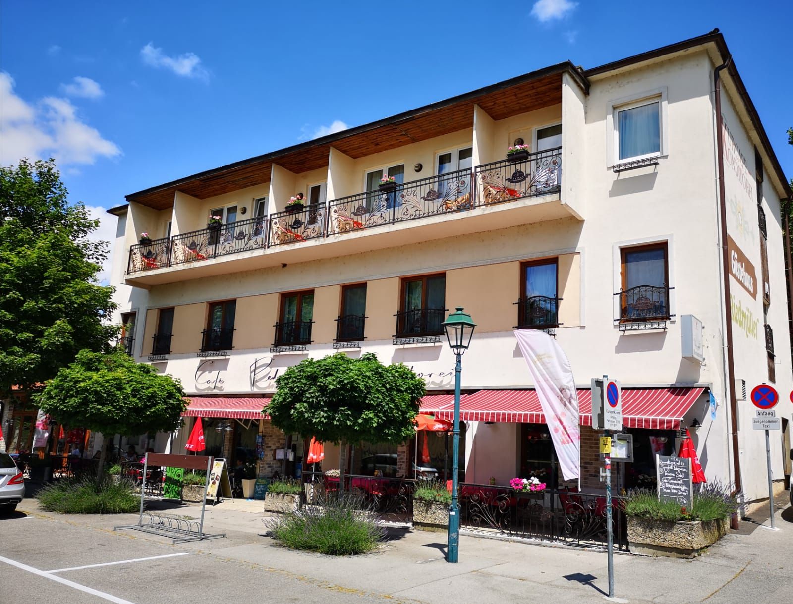 Außenansicht eines Cafés mit Balkon und Markise, umgeben von Bäumen und Straßenschildern.