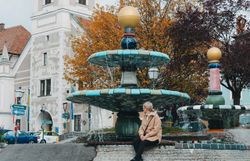 Der Herbst hüllt den Platz in sanfte Farben, während das Wasser des kunstvollen Brunnens sanft plätschert. Eine ruhige Atmosphäre lädt dazu ein, die Schönheit der Umgebung zu genießen und die kreative Architektur zu bewundern.