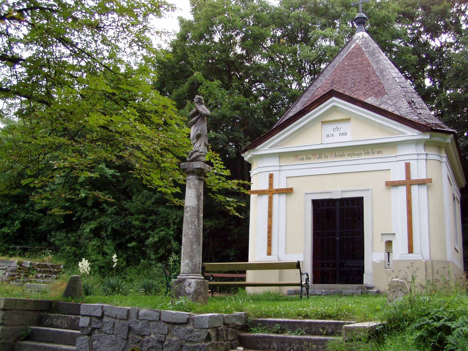 Kapelle mit Statue und Kreuz in einem grünen Park.