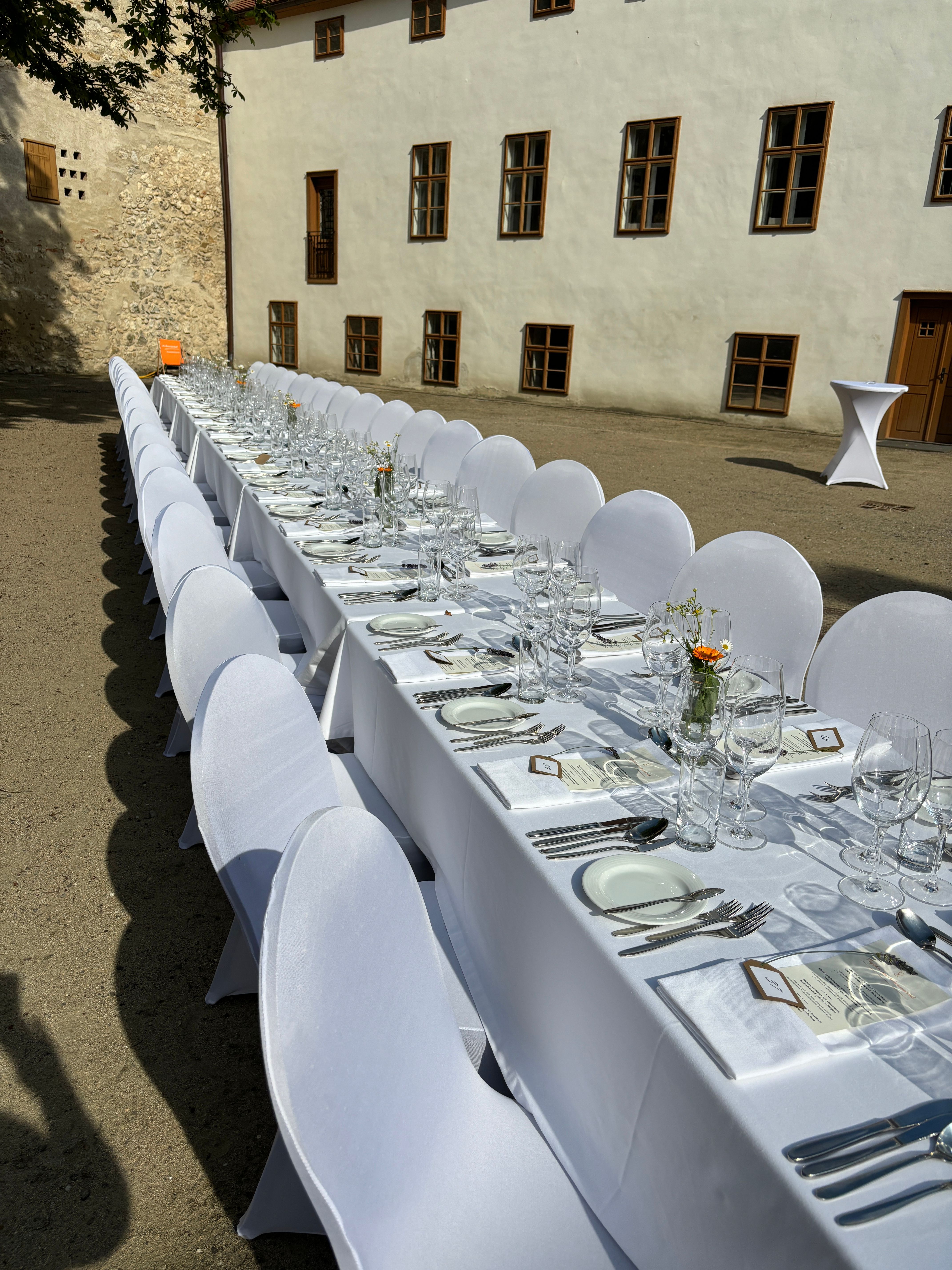 Lange Tafel im Freien mit weißer Tischdecke und Gedecken vor einem historischen Gebäude.