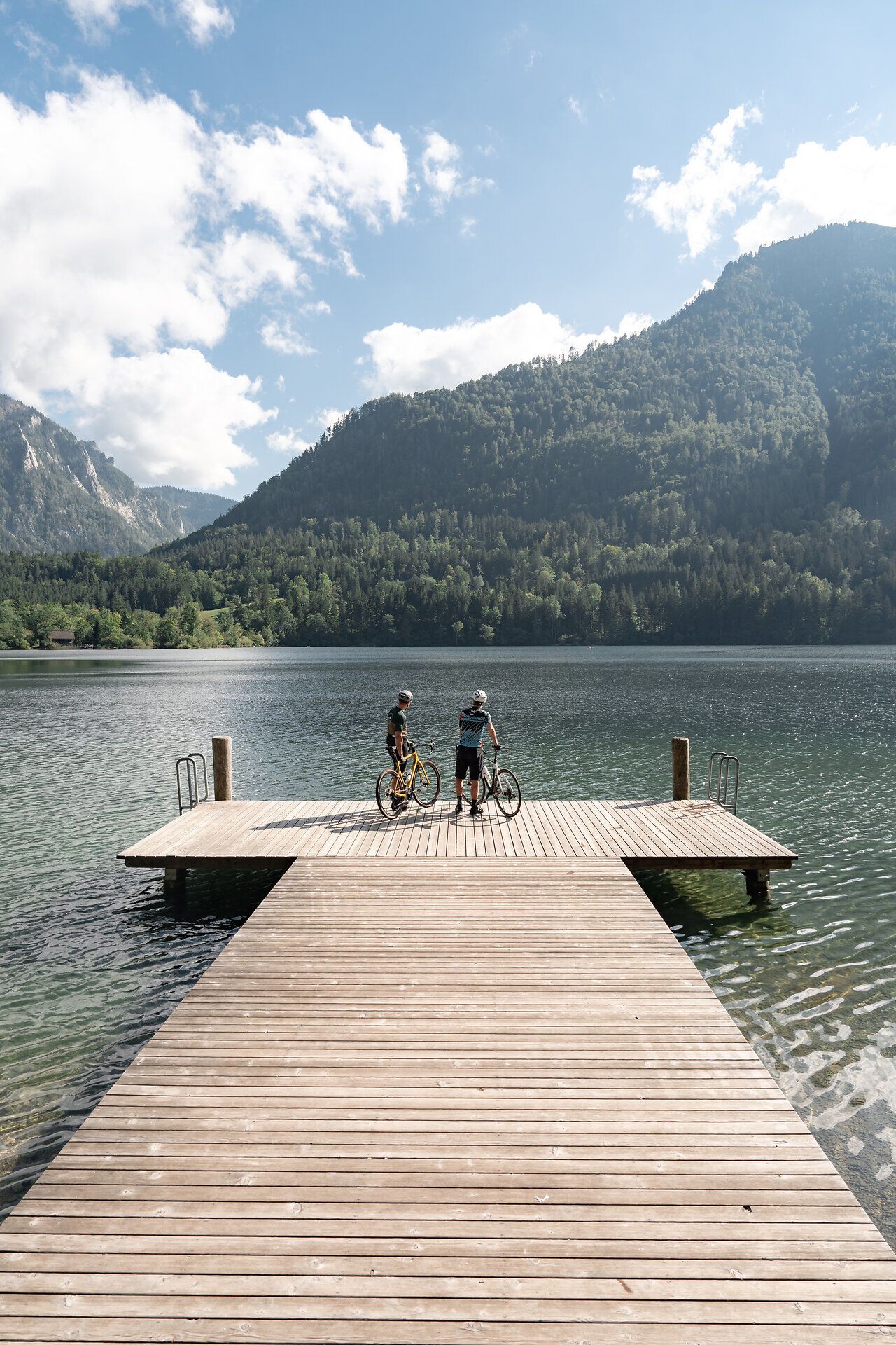 Die sanften Wellen des Sees spiegeln die majestätischen Berge wider, während zwei Radfahrer auf dem Holzsteg innehalten und die atemberaubende Aussicht genießen. Umgeben von üppigem Grün und der frischen Bergluft, lädt dieser Ort dazu ein, die Seele baumeln zu lassen und die Schönheit der Natur zu erleben.
