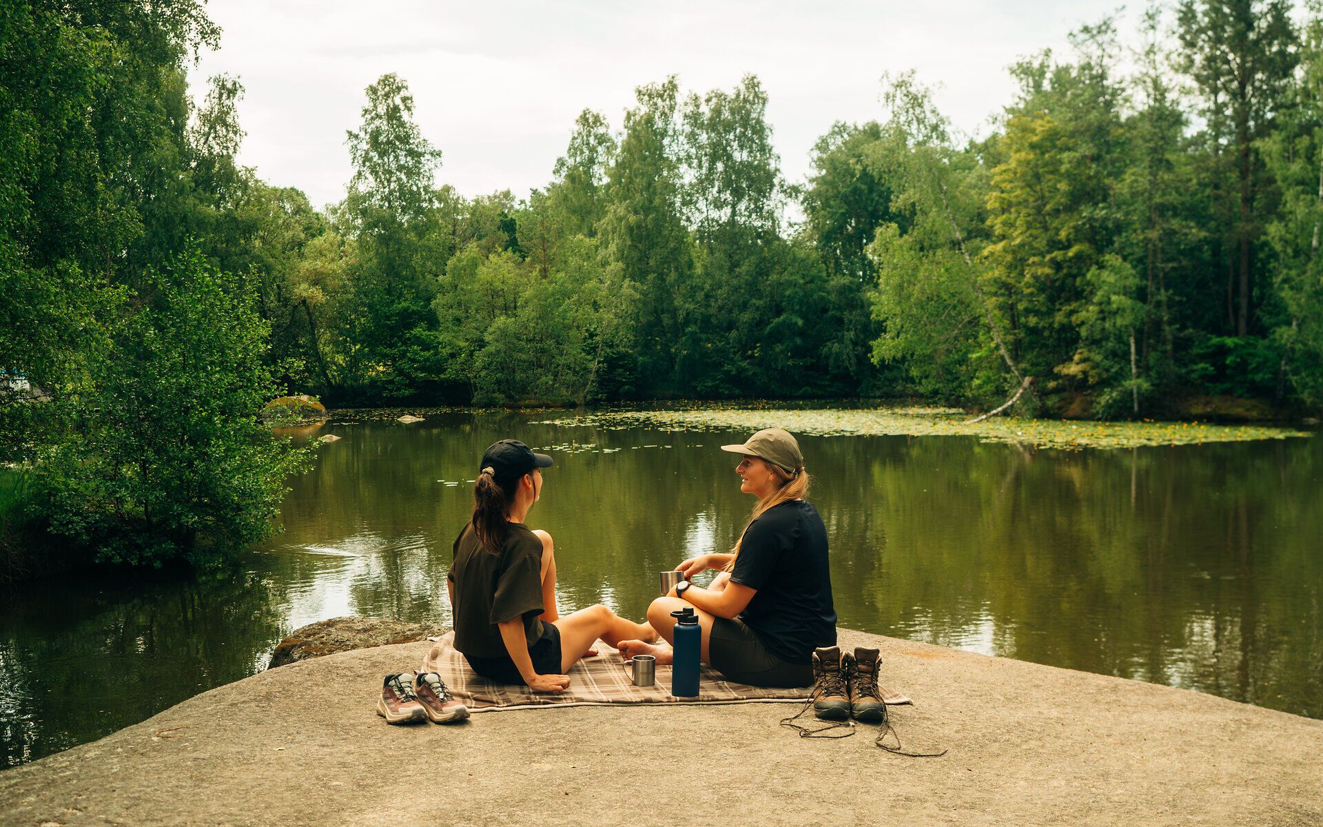 Zwei Frauen sitzen auf einem Stein in der Blockheide, umgeben von Wald und Wasser und machen eine Pause vom Wandern