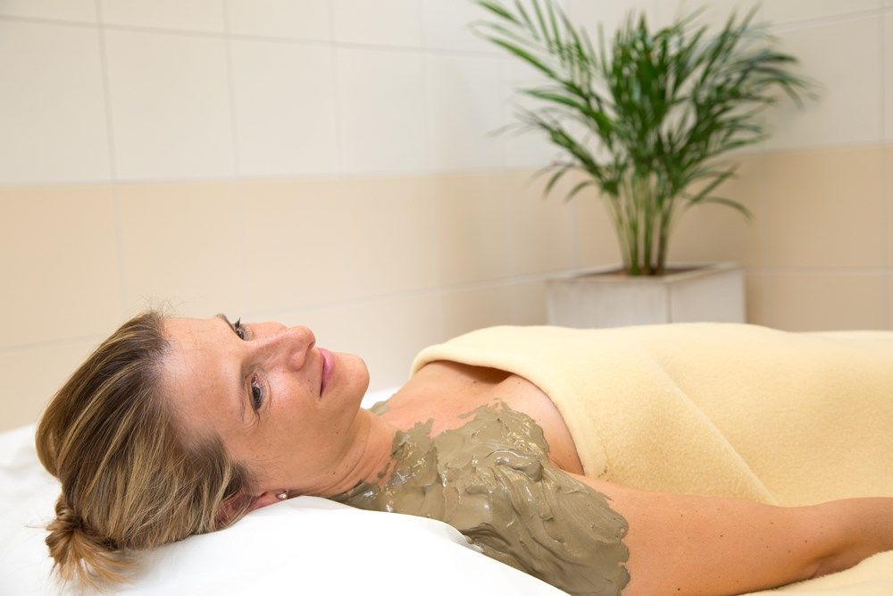 Frau entspannt mit Schwefelschlammpackung auf der Schulter, eingewickelt in eine Decke, im Wellnessbereich mit Pflanze im Hintergrund.