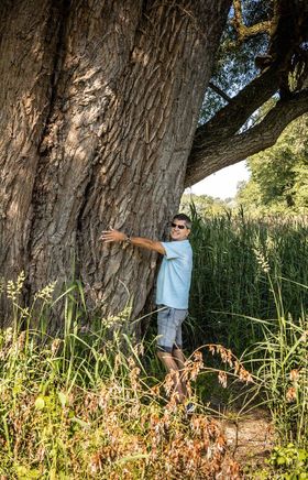 Umgeben von üppigem Grün und der majestätischen Präsenz eines alten Baumes, strahlt die Szenerie eine friedliche Sommeratmosphäre aus. Ein Abenteurer umarmt den Stamm, während die sanften Wellen des Wassers in der Nähe leise plätschern. Diese idyllische Umgebung lädt dazu ein, die Natur in vollen Zügen zu genießen.